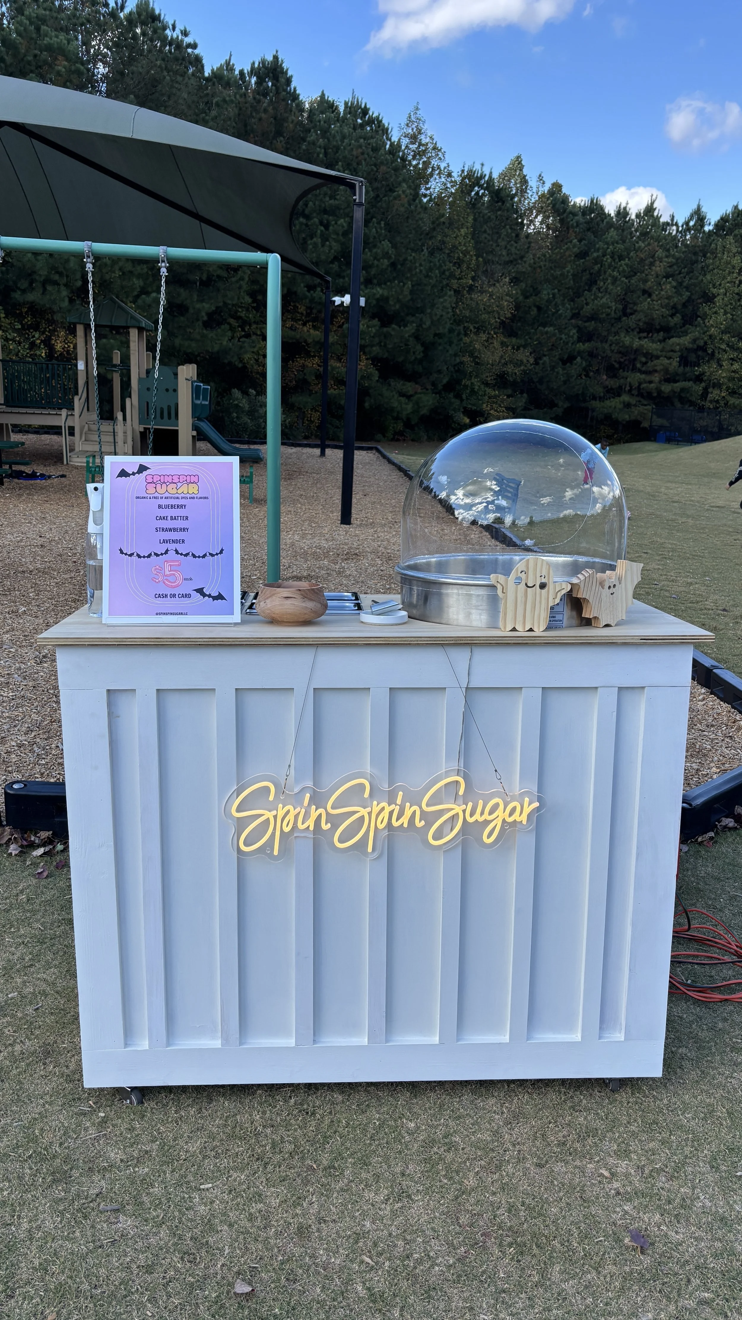A cotton candy stand outdoors in a park with a sign that reads 'SpinSpinSugar.' The stand has a yellow neon sign and offers candy flavors such as blueberry, cake batter, strawberry, and lavender for $5. The setup includes a cotton candy machine, a wo