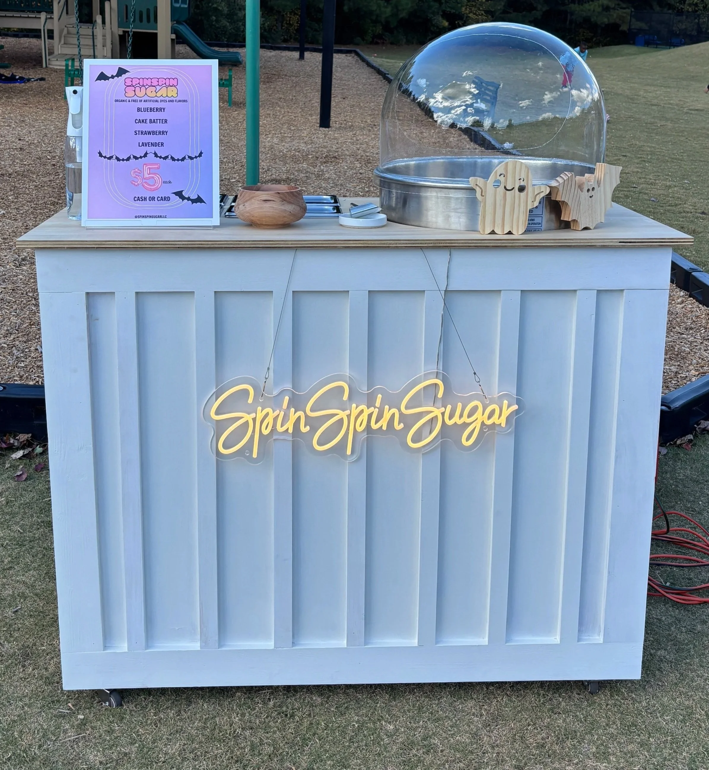 A white wooden vendor cart with a neon sign reading 'Spin Spin Sugar'. On top, there is a sign offering flavored spun sugar for $5, a wooden container, and a clear plastic dome. There are two wooden decorative bats on the cart. The background includes a playground and a grassy field.