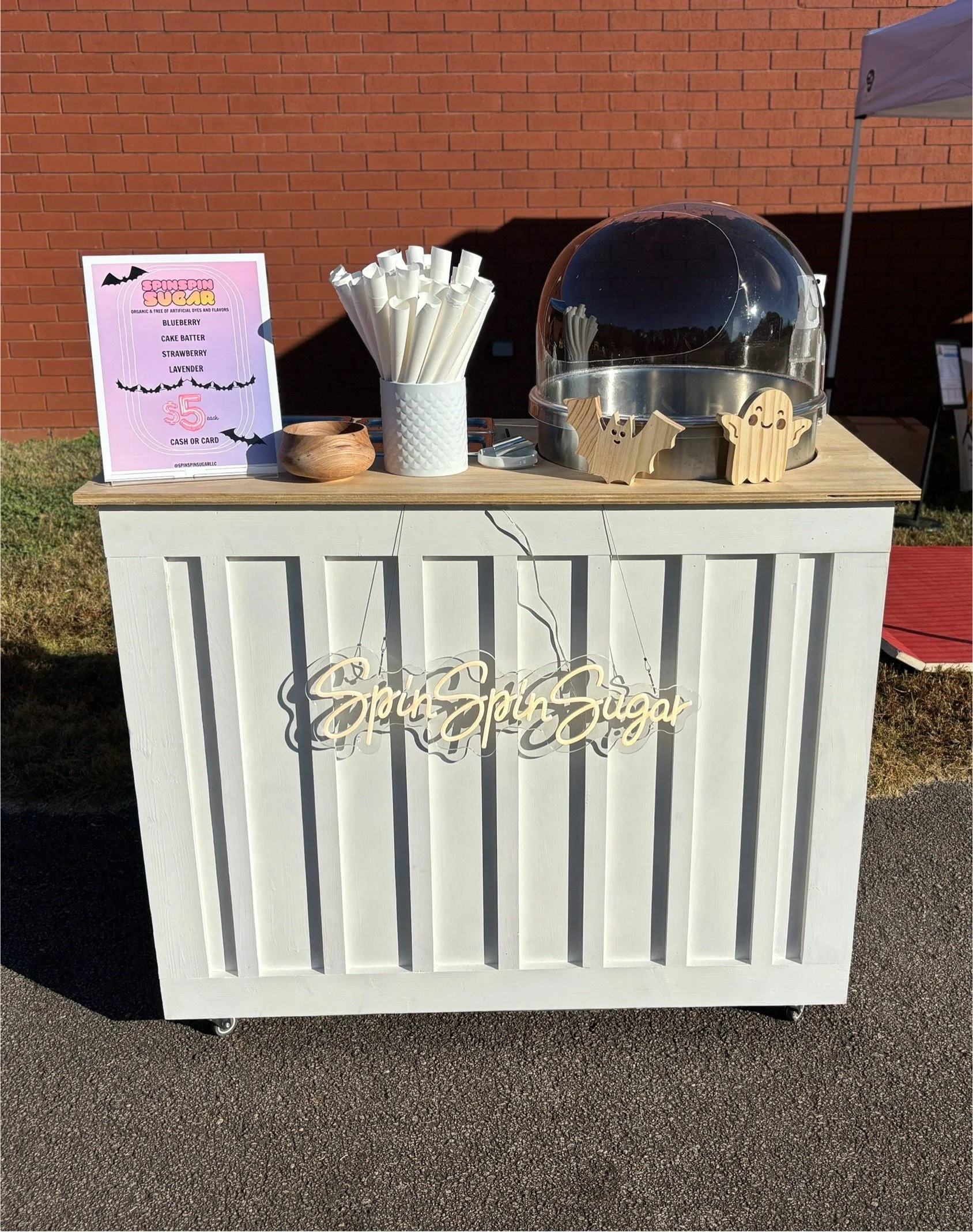 A cotton candy stand with a white wooden base and a light wood top, featuring a neon sign that reads 'Spin Spin Sugar'. There are white paper twist cups, a wooden bowl, Halloween-themed wood decorations, a large metallic fruit stand cover, and a pink