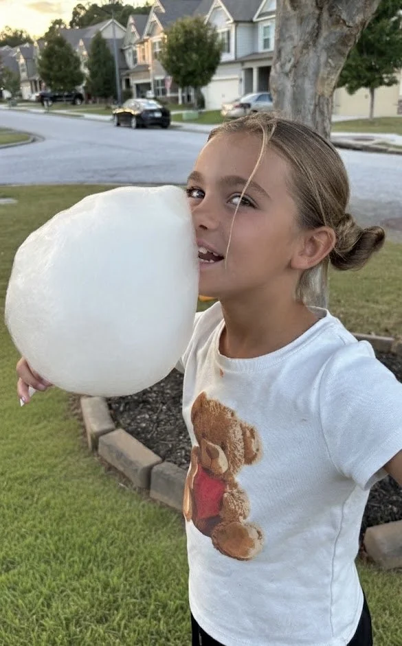 A girl with braided hair holding a large piece of cotton candy outside near a sidewalk and houses.