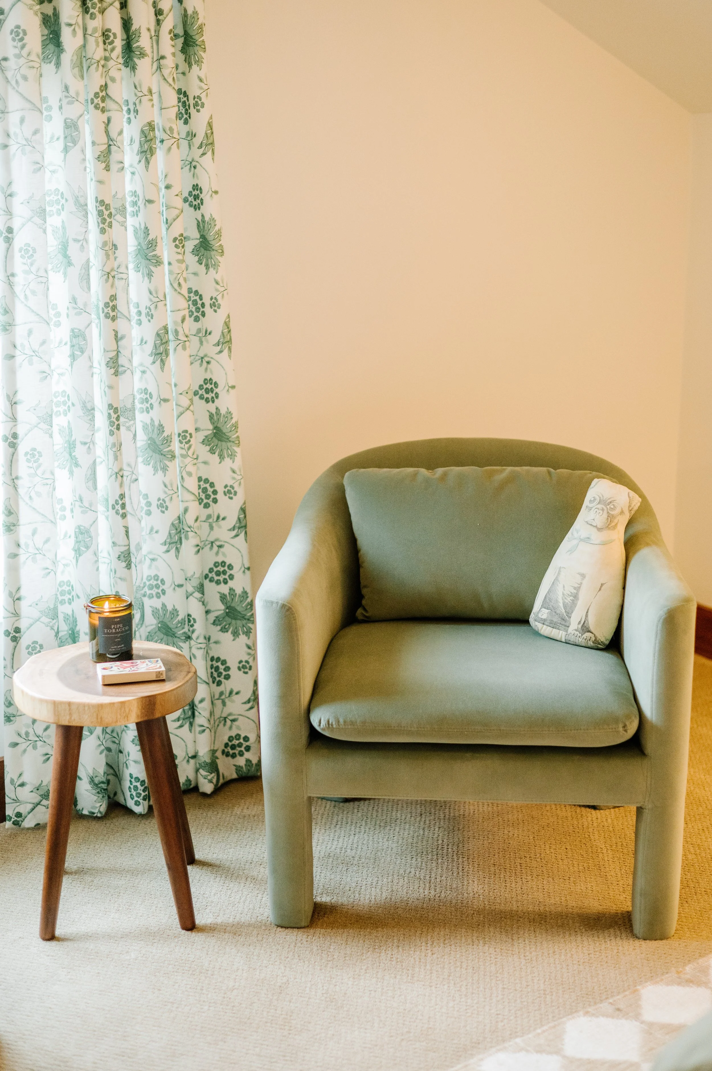 A green armchair with a matching cushion and a decorative pillow featuring a face, next to a small wooden side table holding a candle and a box of matches, with leafy patterned curtains in the background.