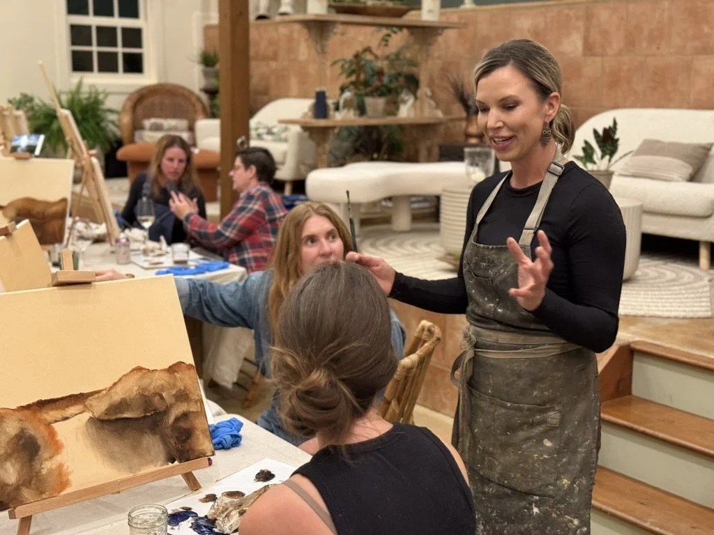 An art instructor explaining painting techniques to students at an art class in a cozy, well-lit room with couches and plants.