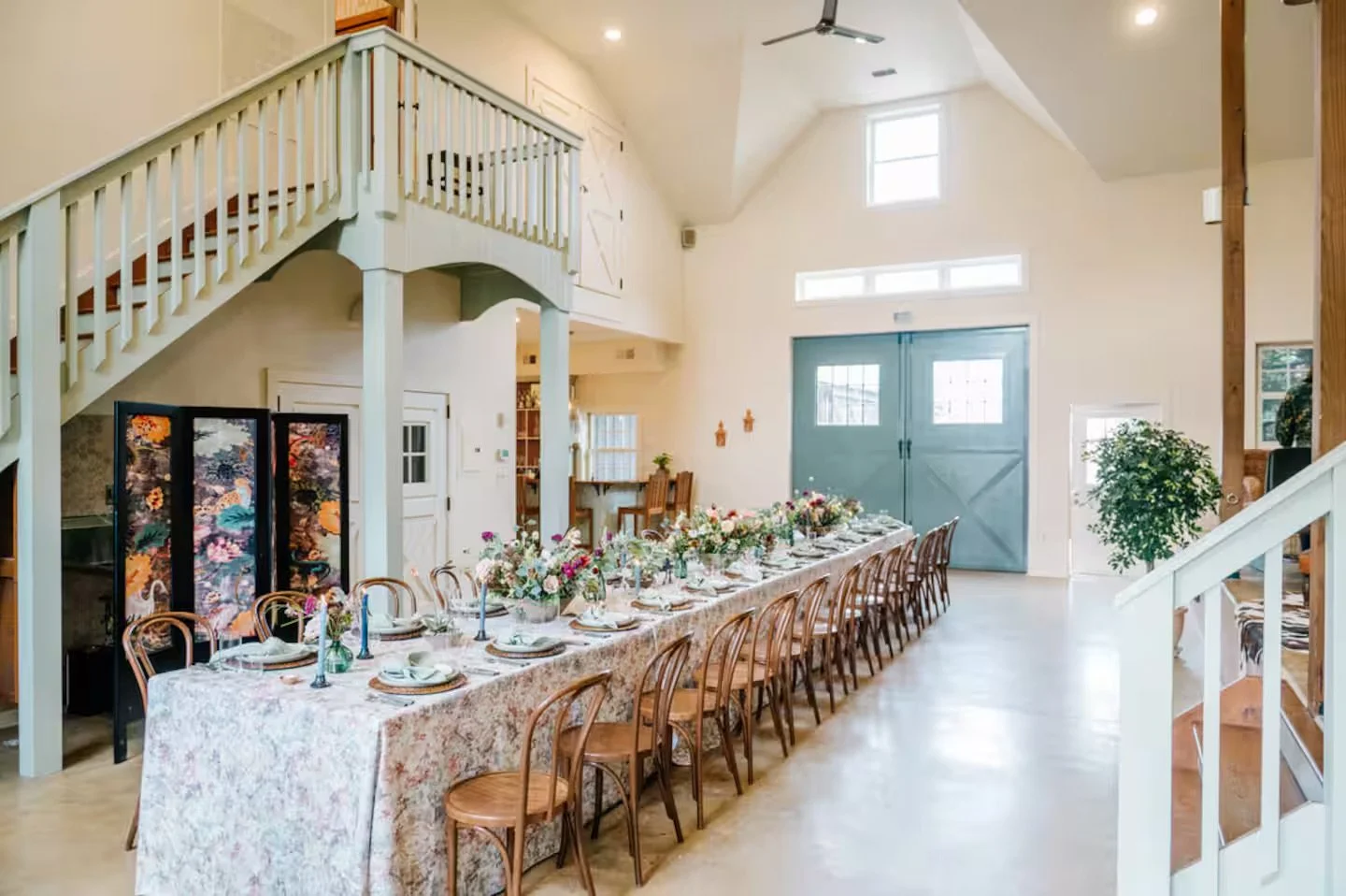 Long dining table with floral tablecloth and floral centerpieces, set with plates, napkins, and candles, in a spacious, well-lit room with large blue double doors.