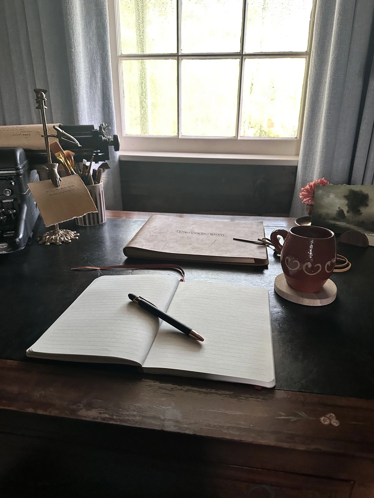A desk with an open notebook and pen, a closed notebook titled "The Tales Cabin," a red mug on a coaster, and a window with curtains in the background.