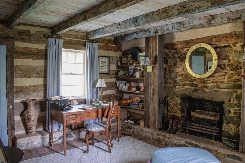 A rustic room with wooden beams, a window with light blue curtains, a wooden desk with a typewriter and books, and a stone fireplace with a mirror and boots on the hearth.