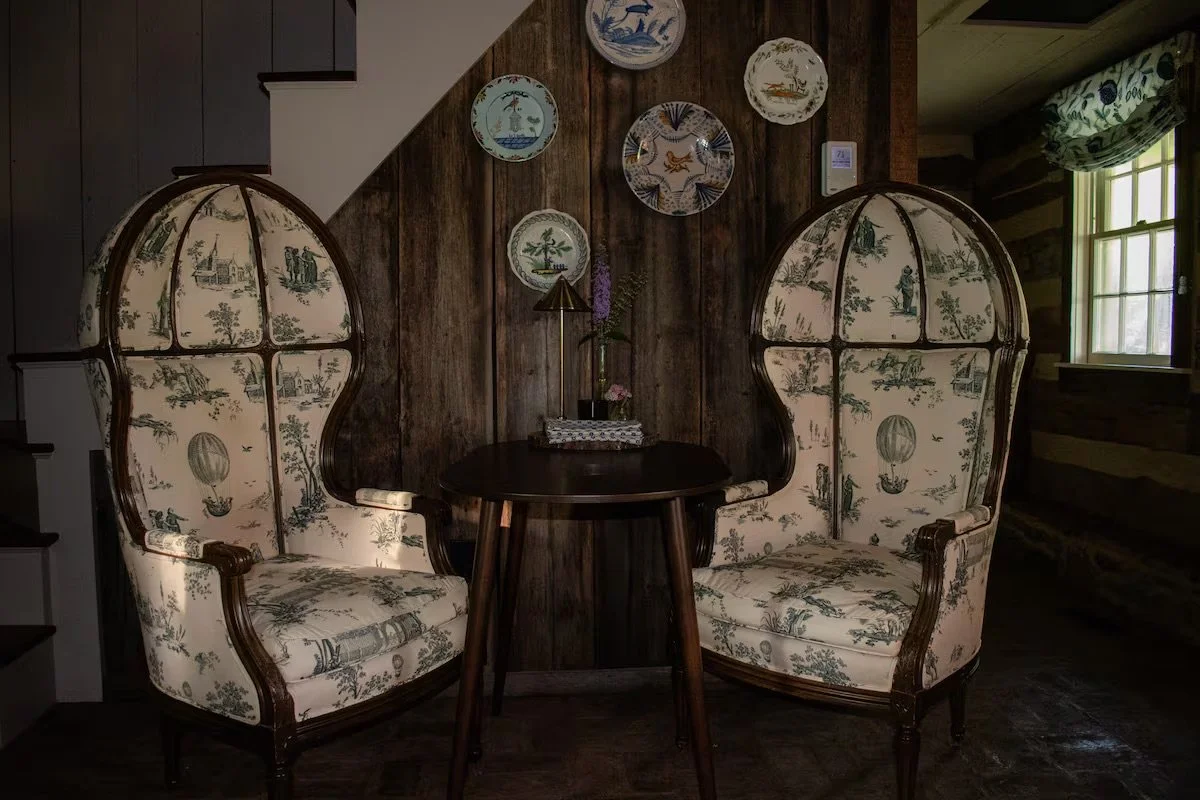 A cozy corner with two high-backed, vintage upholstered armchairs with botanical and scenic patterns, facing each other across a small dark wooden round table, which holds a small decorative tray with flowers and a brass lamp. The background features