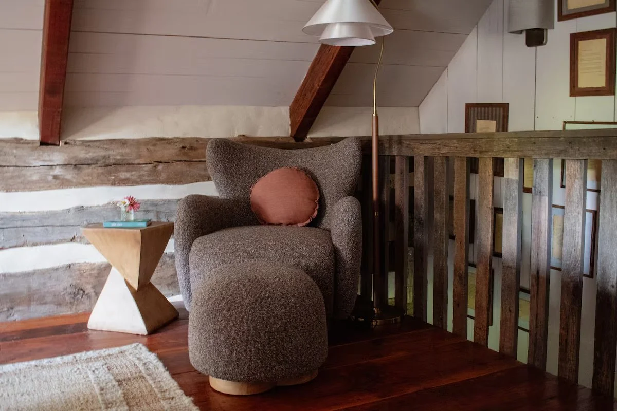 Cozy corner with a gray upholstered armchair, a matching footstool, a round rust-colored pillow, a small wooden side table with a flower in a vase and a book, a floor lamp with a white shade, wooden beams, and framed pictures on the wall in a rustic 