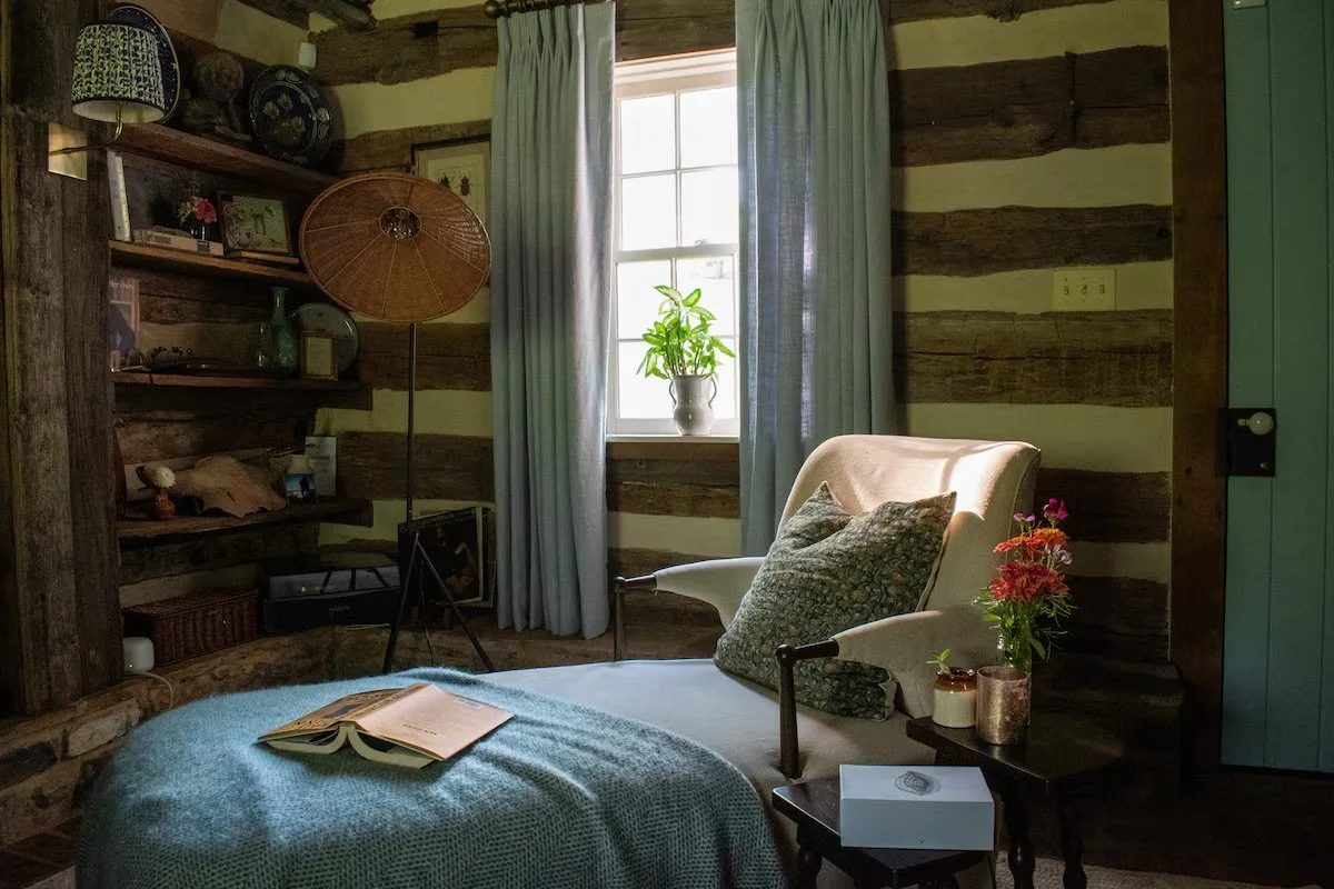 Cozy rustic bedroom with wooden logs on the wall, a bed with an open book, an armchair with pillows, a window with curtains and a potted plant, and a side table with flowers and candles.