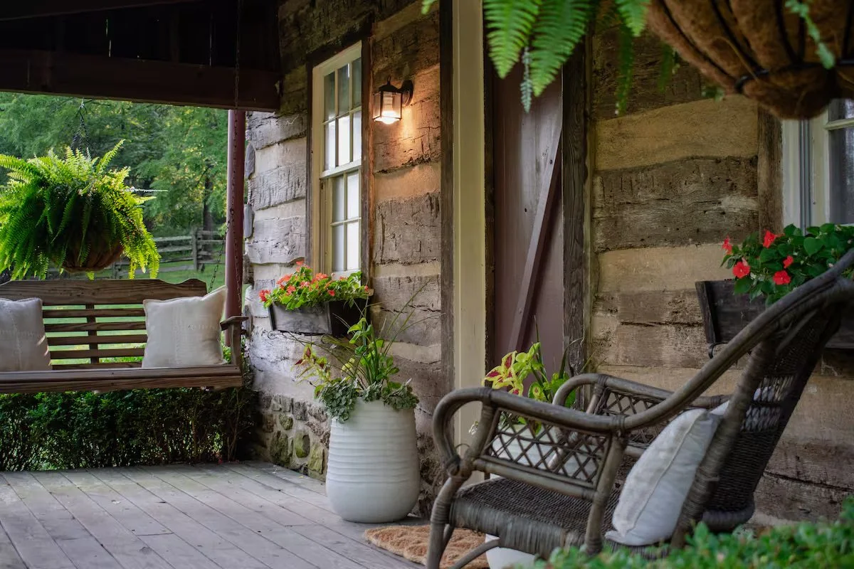 A cozy outdoor porch with a wooden swing with white pillows, a wicker chair with a pillow, and potted plants hanging and placed beside the wall of a rustic stone and wood cabin.