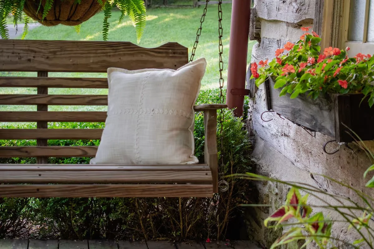 A wooden porch swing with a white pillow, hanging near a stone wall with window boxes filled with pink and orange flowers, surrounded by greenery.
