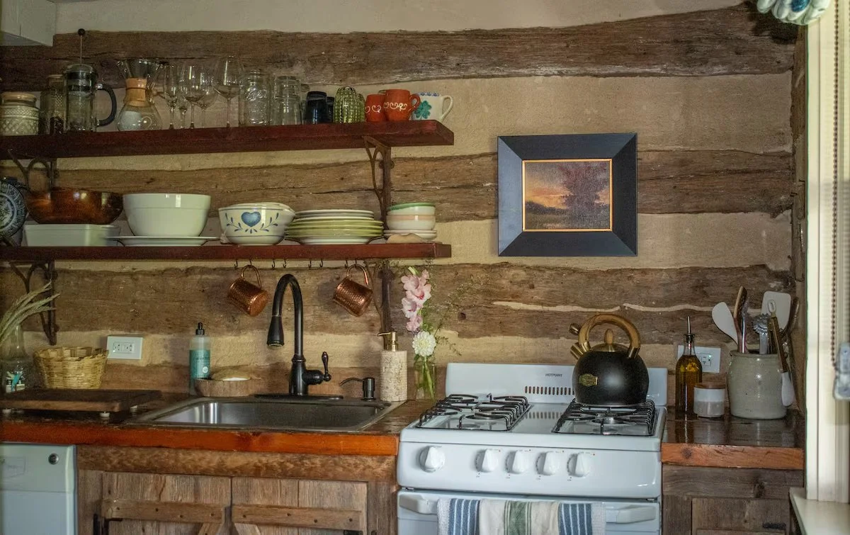 A rustic kitchen wall with wooden beams and shelves holding dishes, glasses, cups, and kitchen utensils. There is a black kettle on the stove, a vase with pink flowers, and various bottles and containers on the countertop.
