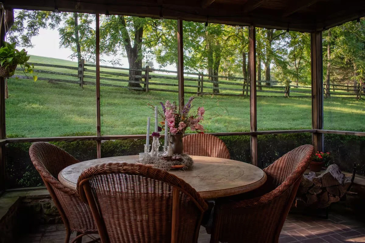 A wooden indoor dining area with a round table and four wicker chairs, decorated with a pink floral centerpiece, candles, and natural light from large screened windows overlooking a green yard with trees and a wooden fence.