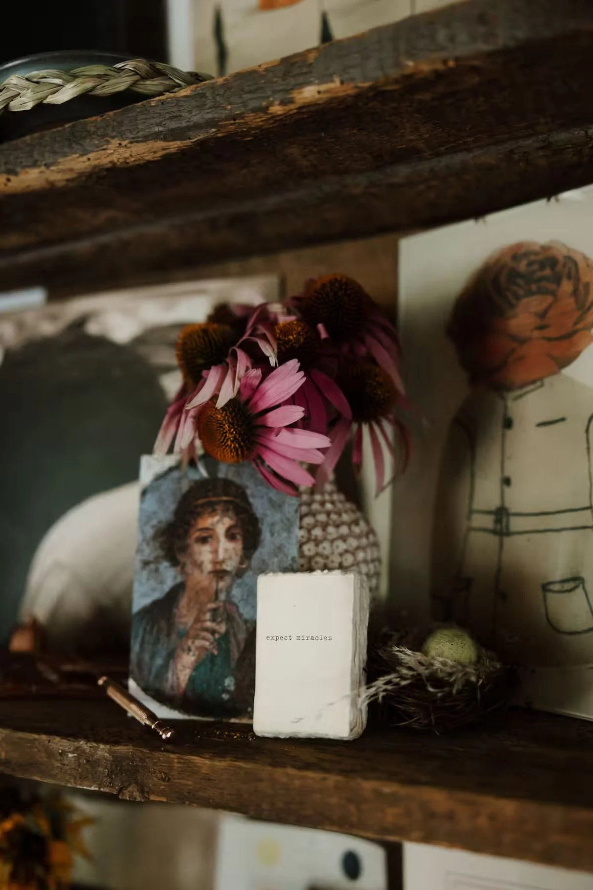 A small decorative shelf with a vintage painting of a woman drinking from a straw, a bunch of pink and brown flowers in a container, a small marble block with the words 'expect miracles' written on it, and various other decorative objects and drawing