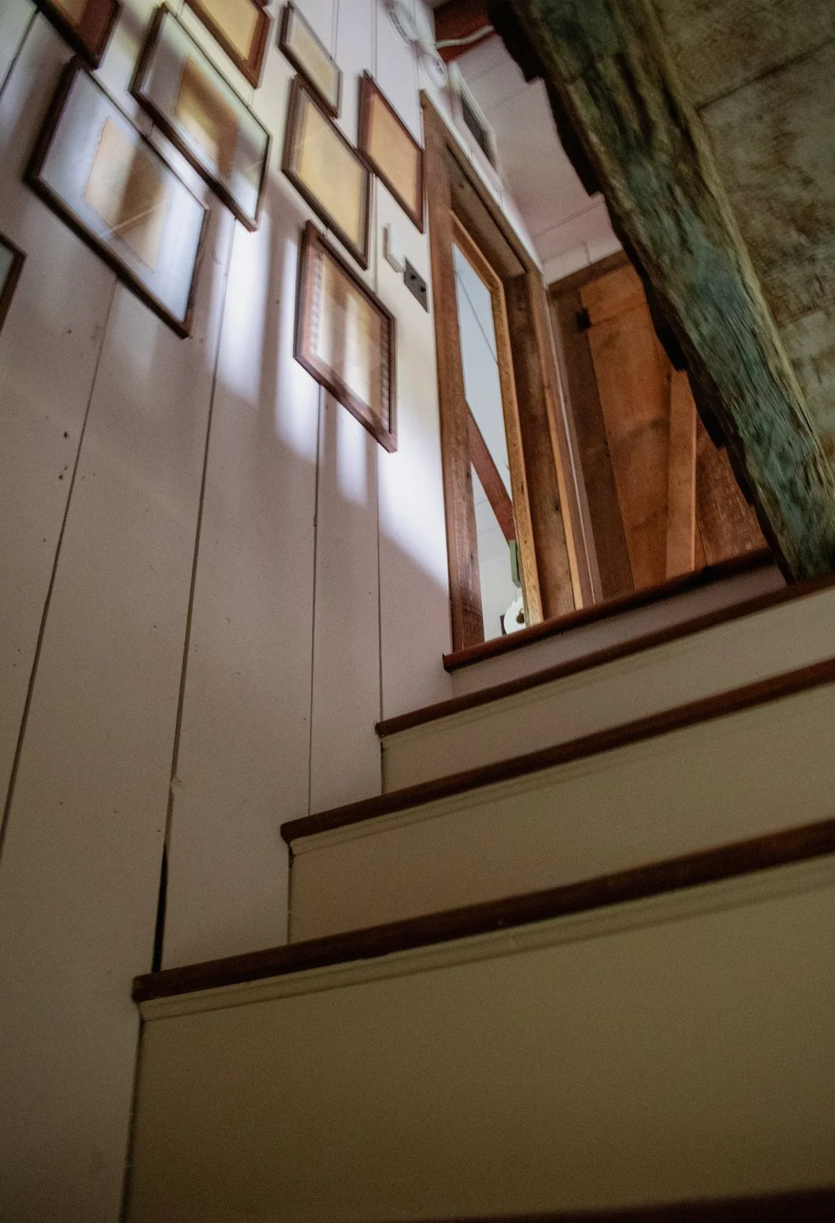 Inside view of a staircase with beige steps and brown edges, leading up to a hallway with framed pictures on the wall. The photo is taken from the bottom of the stairs looking up.