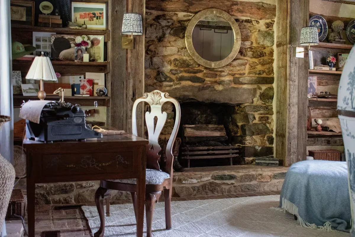 A cozy living room with a stone fireplace, a white carved chair, a wooden desk with a vintage typewriter, bookshelves filled with books and decorative items, and various lamps providing warm lighting. There is also a mirror above the fireplace and a 
