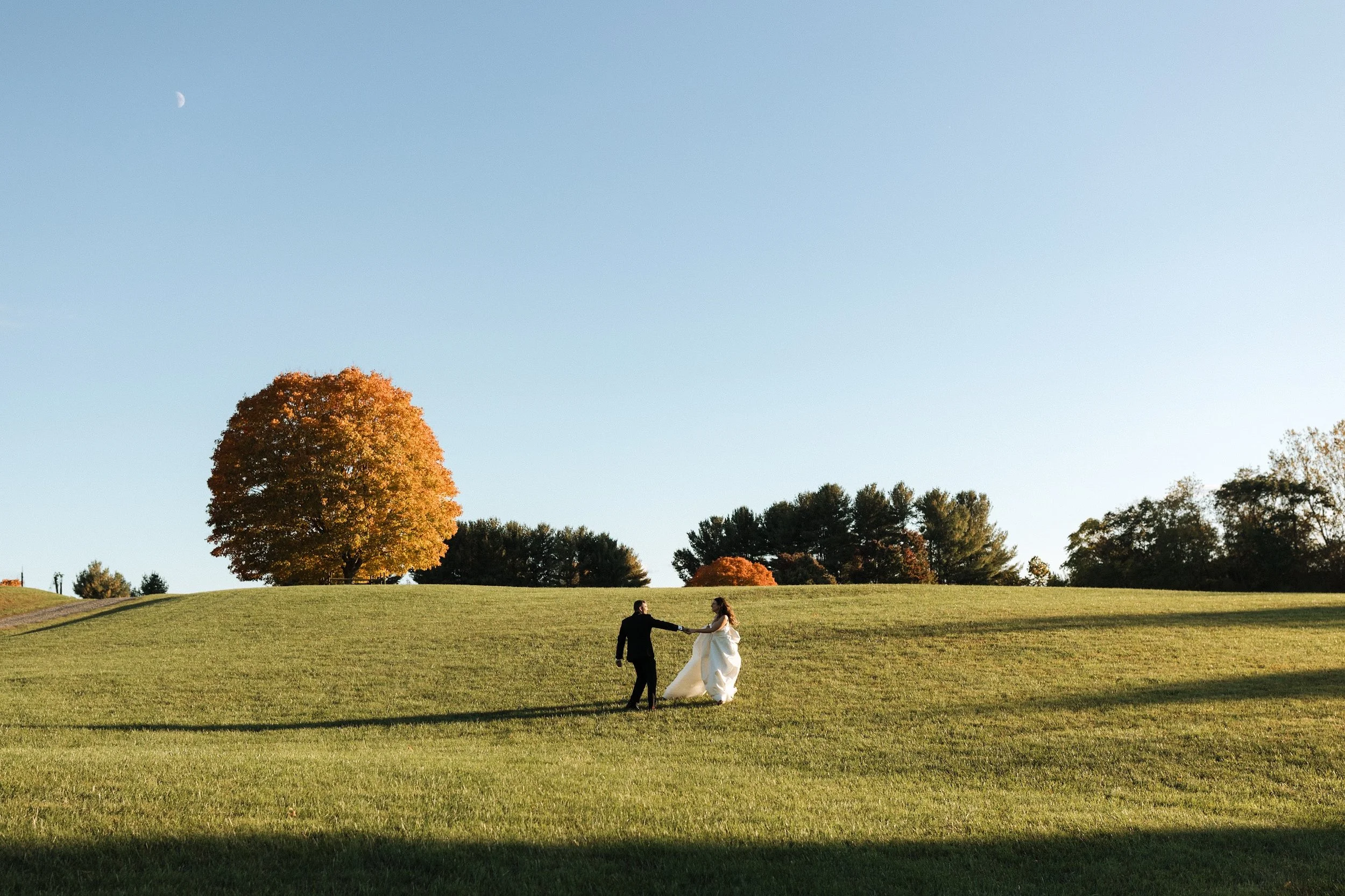 A bride and groom holding hands and dancing on a grassy hill under a clear blue sky with a crescent moon, trees with autumn foliage in the background.