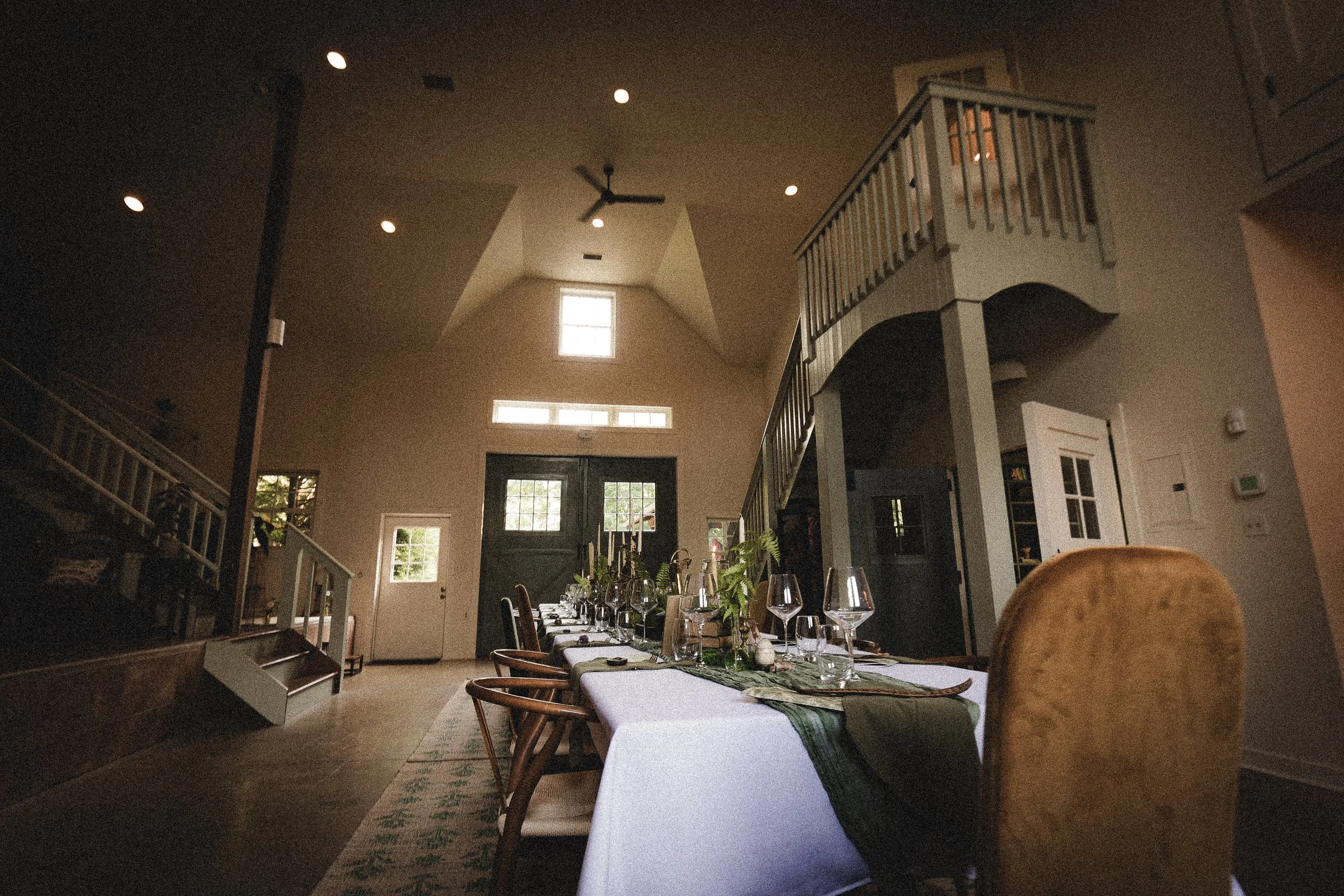 Interior of a dining room with a long table set with glasses and greenery, high ceiling, staircase on the right, and a door at the back.