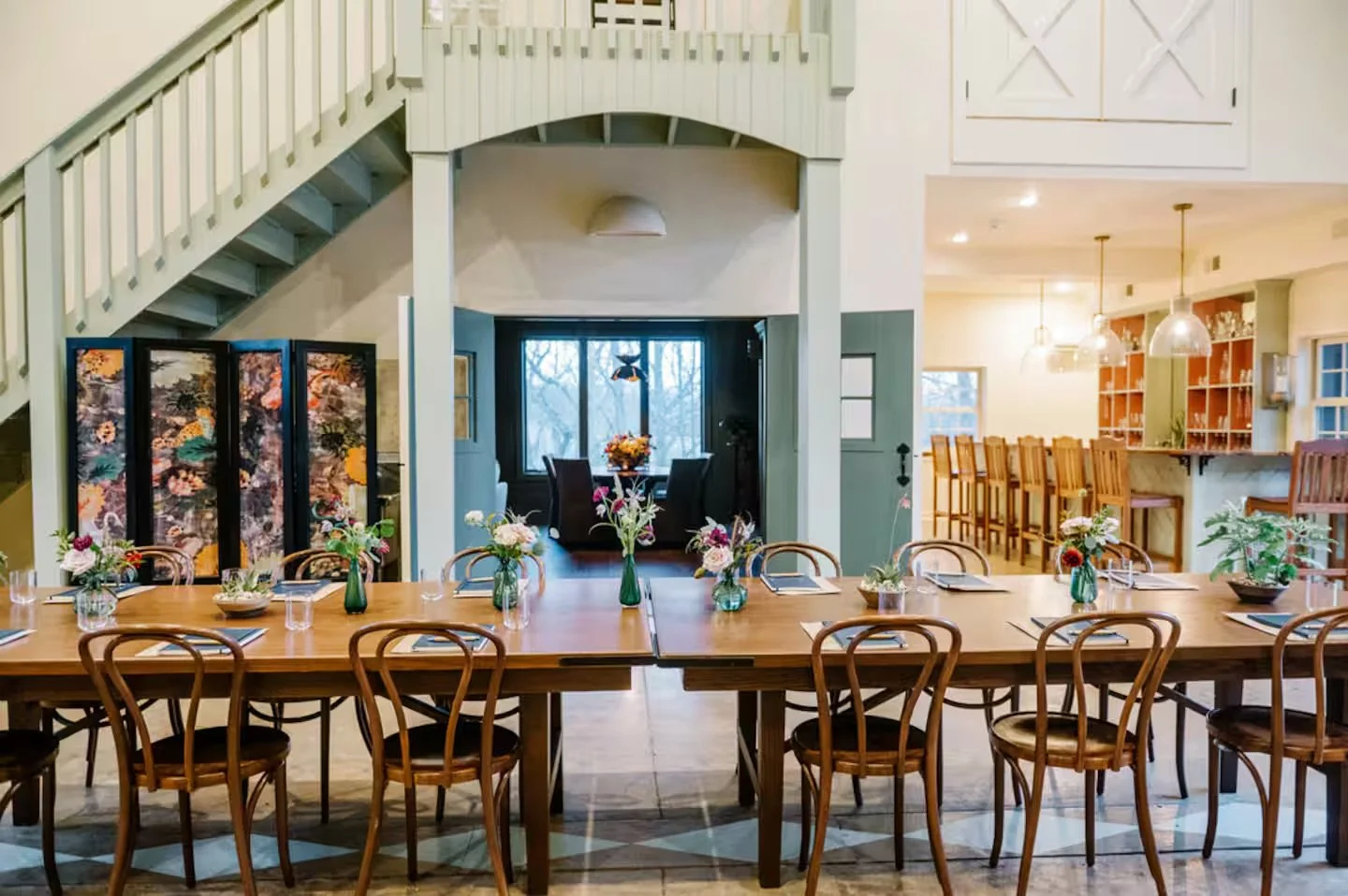 A cozy dining area with a long wooden table set with small flower arrangements, surrounded by wooden chairs, in front of a room with a bay window, and an open kitchen with bar stools, pendant lights, and shelves.
