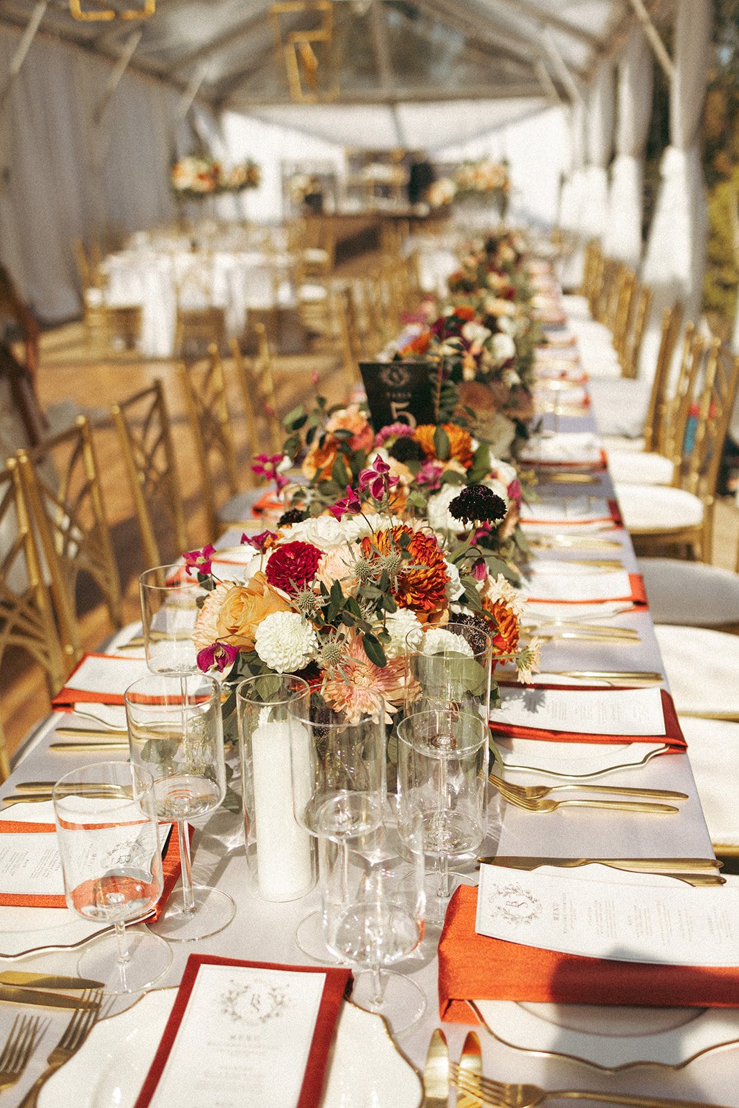 Decorated long banquet table with floral centerpieces, gold cutlery, wine glasses, and place settings, set inside a tent for an event.