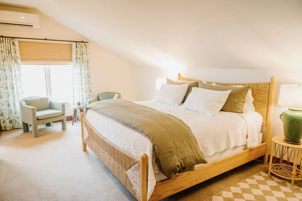 A cozy attic bedroom with a large bed, light-colored bedding, and decorative pillows. There's a nightstand with a green lamp, a sitting area near a window with floral curtains, and two chairs with a small table between them.