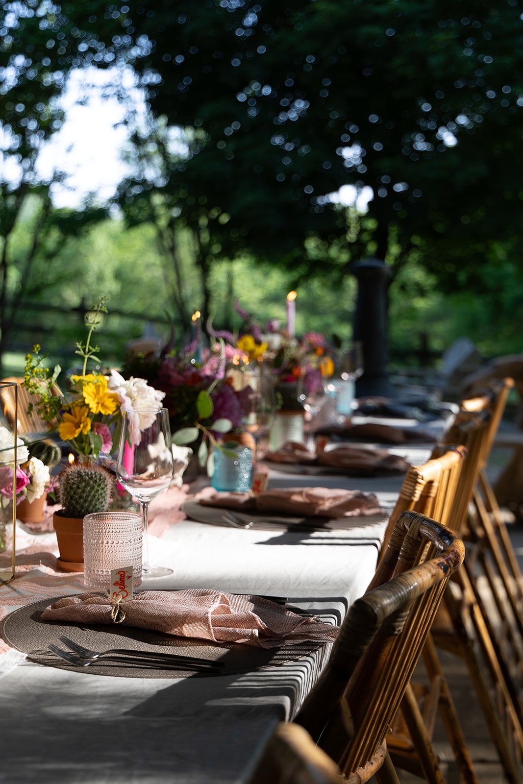 A long outdoor dining table set with plates, napkins, glasses, and centerpieces of flowers and succulents, surrounded by wooden chairs, in a shaded garden area.