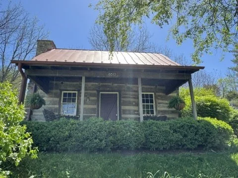 A rustic log cabin with a metal roof, front porch, and surrounding greenery, under a clear blue sky.