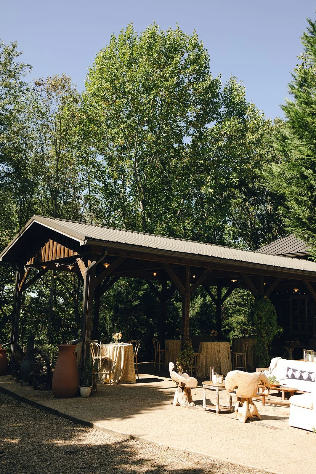 Outdoor patio with tables and chairs under a wooden pavilion, surrounded by trees and potted plants.