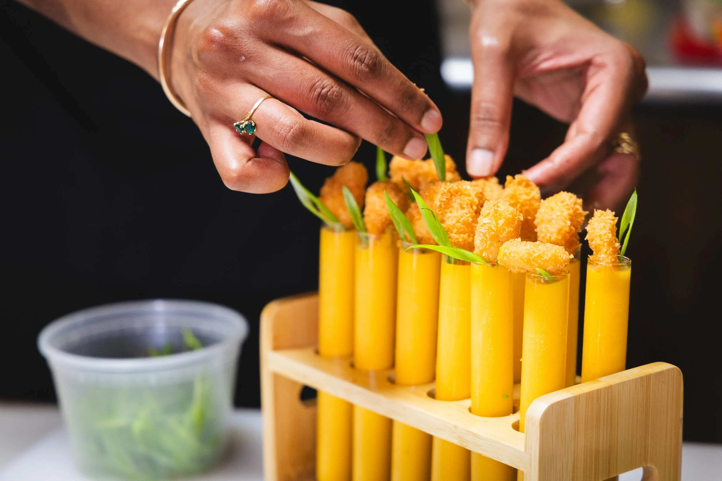 Hands arranging fried fish sticks and green garnishes in a tall yellow holder on a wooden stand, with a small plastic container of greens in the background.