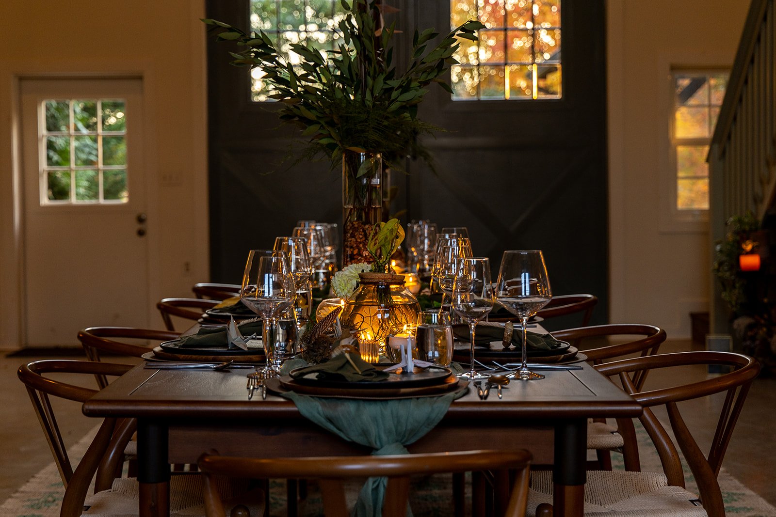 A dining table set for a meal with wine glasses, plates, and cutlery, decorated with a large floral centerpiece and warm candlelight in a cozy room with large windows showing fall foliage outside.