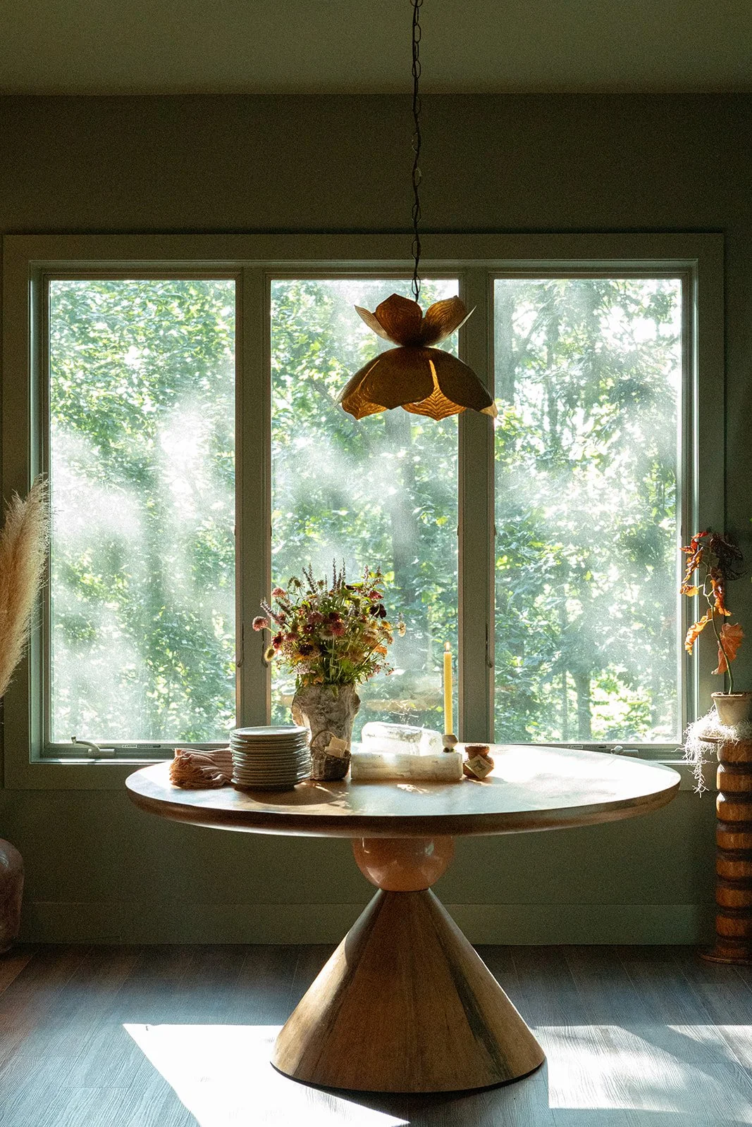 A round wooden dining table with a bundle of plates, a vase of flowers, a candle, and some other items, positioned in front of a large window with trees outside, inside a room with a hanging flower-shaped light fixture.