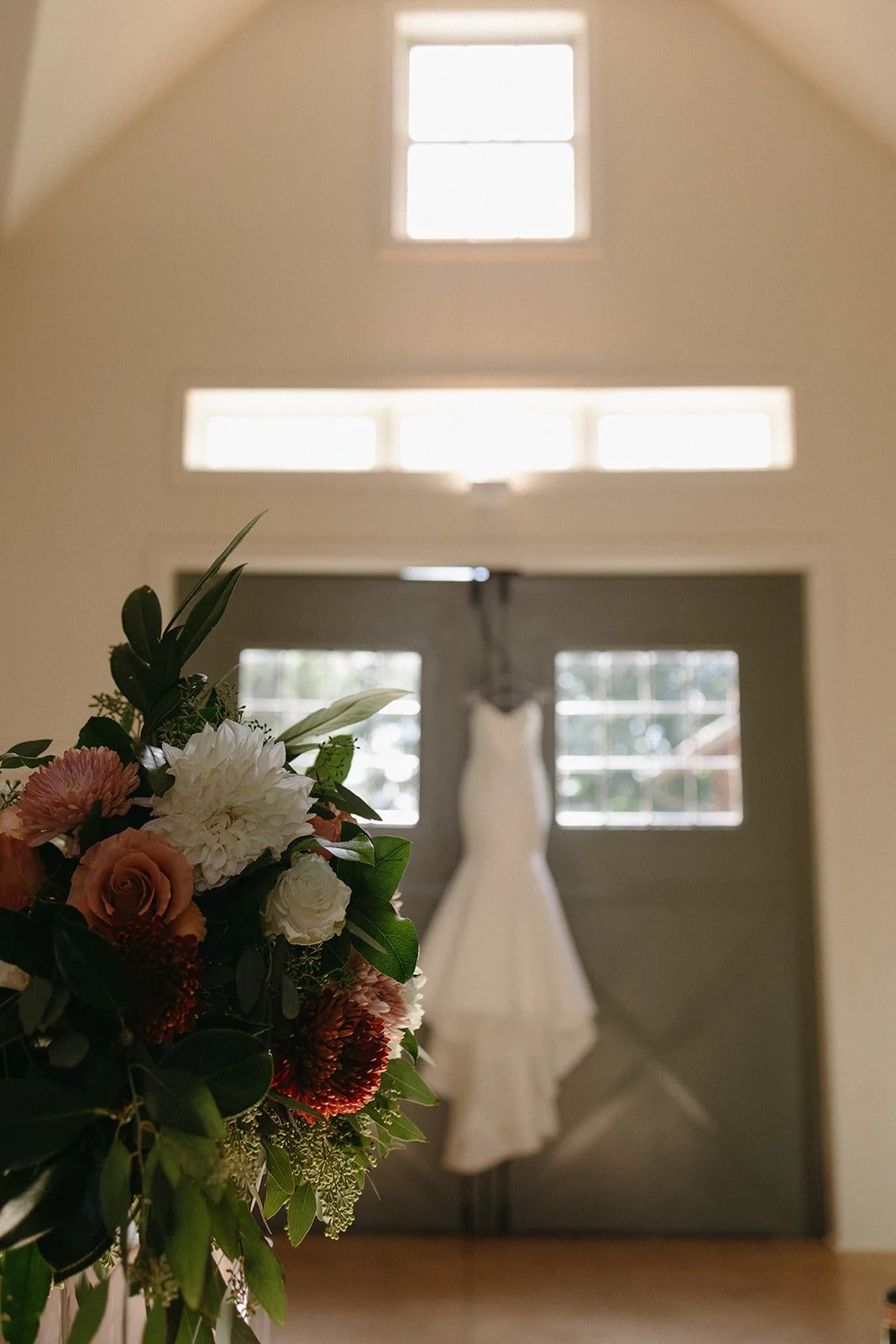 A wedding dress hanging on the back of gray double doors with windows, with a flower arrangement in the foreground.