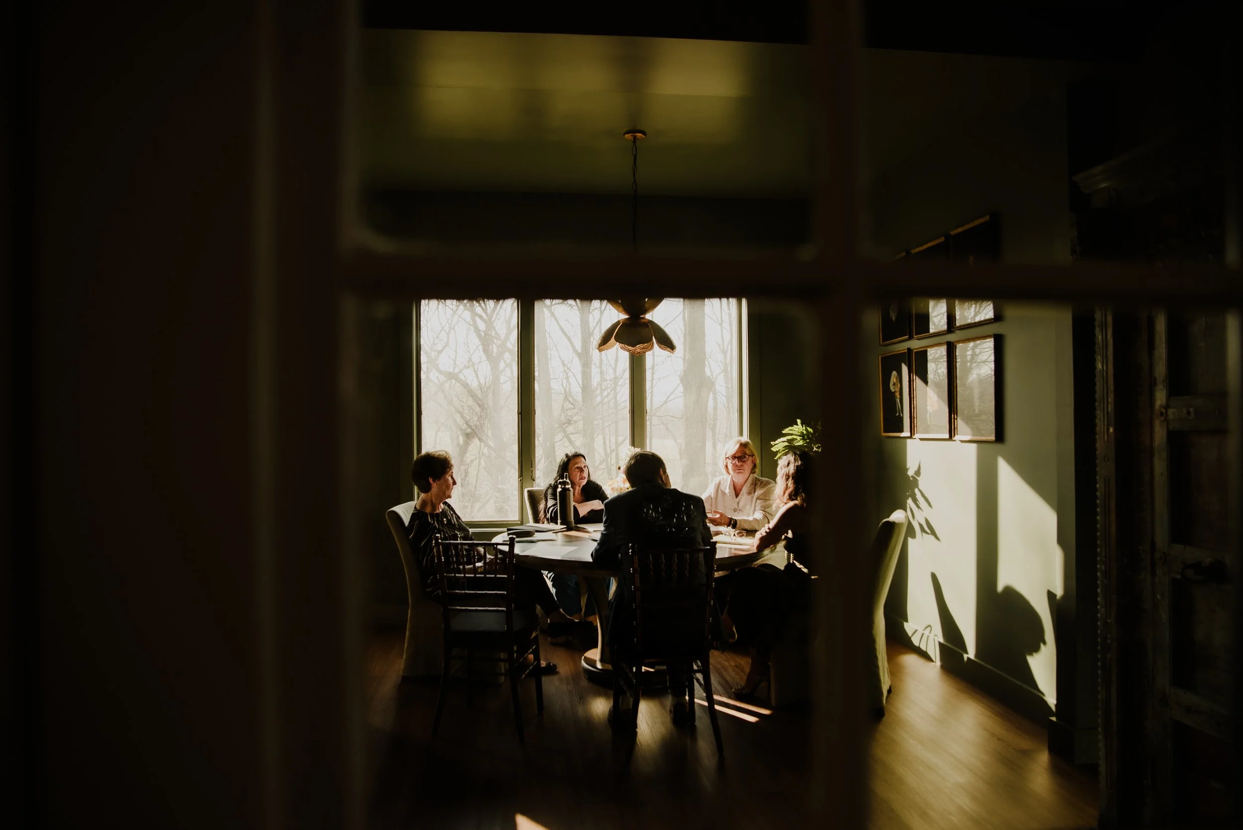 A group of five people sitting around a dining table in a sunlit room with large windows and framed pictures on the wall, viewed from a slightly hidden perspective.