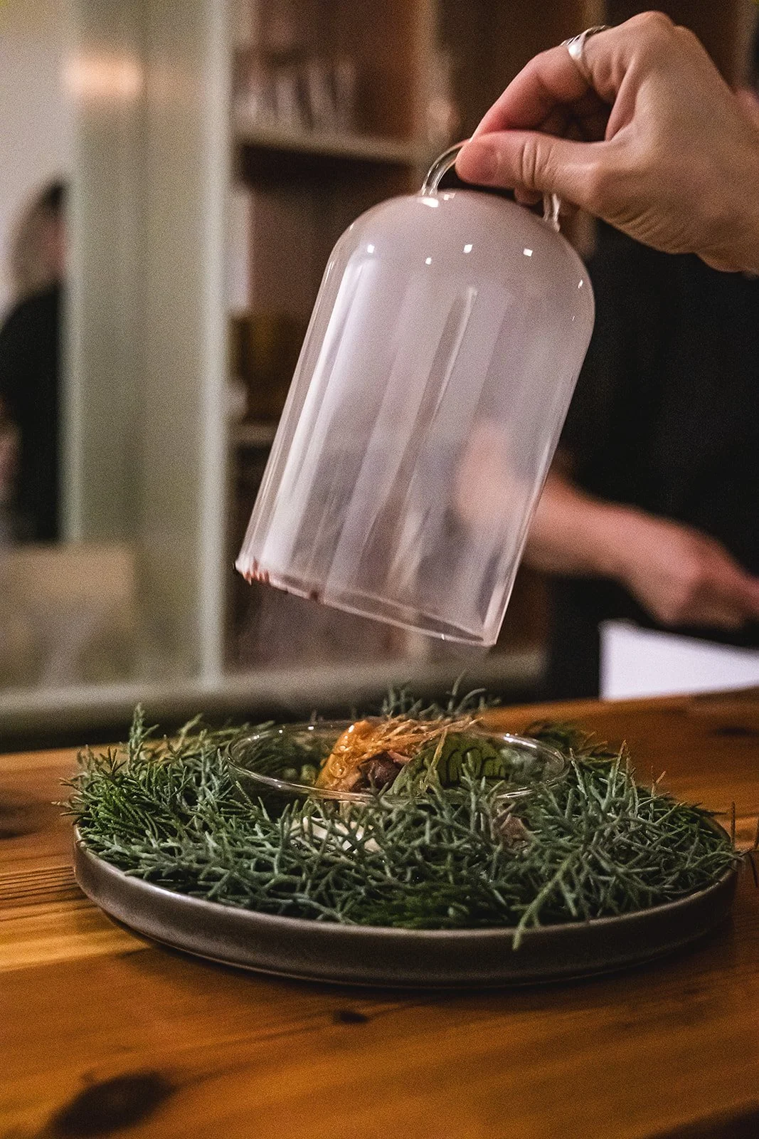 Person sprinkling seasoning onto a dish of food garnished with herbs, on a wooden table.