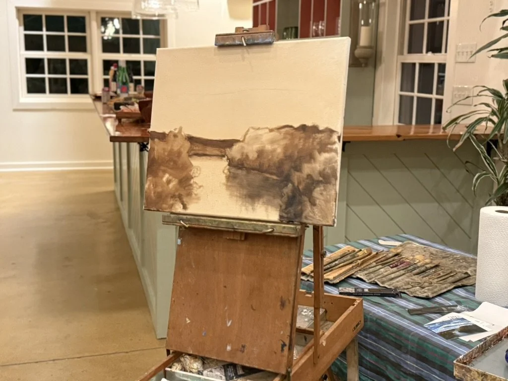 A painting in progress on an easel, depicting a landscape with a body of water and trees, with a kitchen and window in the background.
