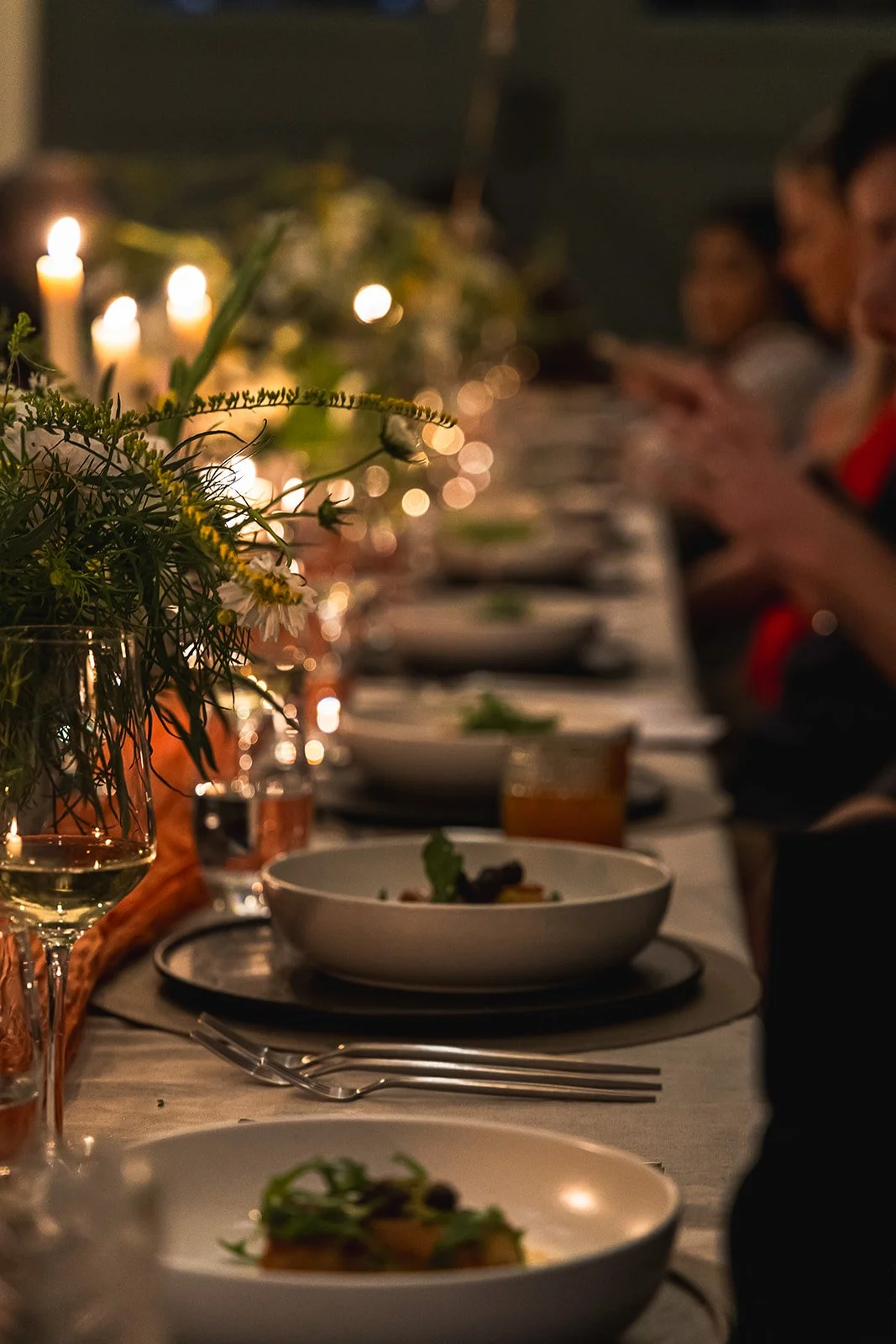 A dinner table set with white plates, wine glasses, flowers, candles, and food, with people dining in the background.