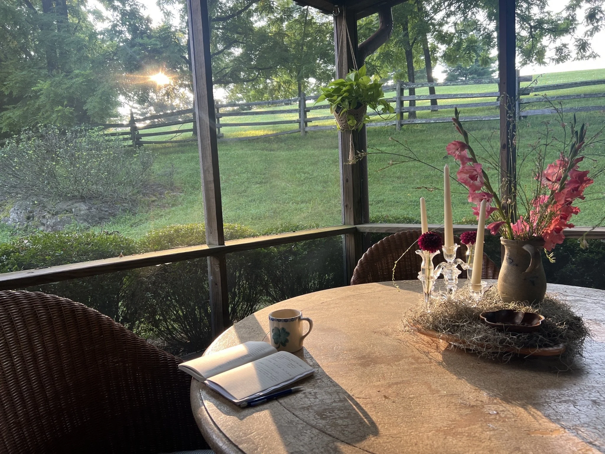 A cozy sunlit screened porch with a round wooden table decorated with a vase of pink flowers, a candelabra, a small bowl, a mug, and an open notebook with a pen, overlooking a lush green lawn and trees outside.