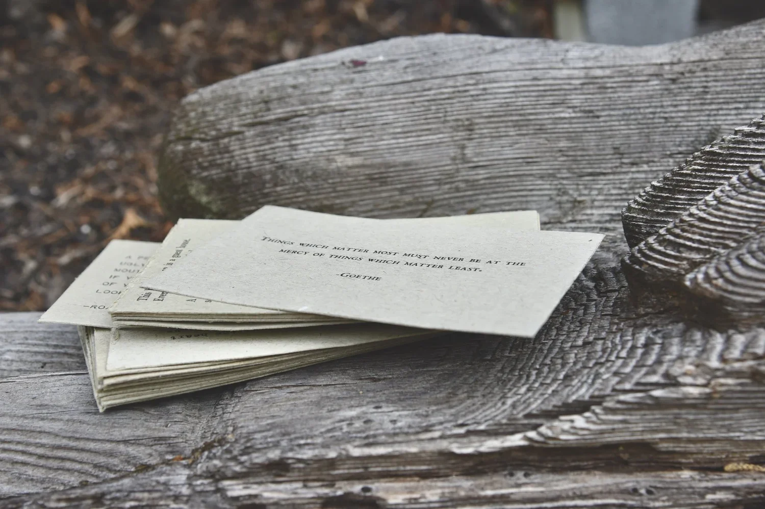 A small stack of paper notes with a quote, resting on a weathered wooden bench outdoors.
