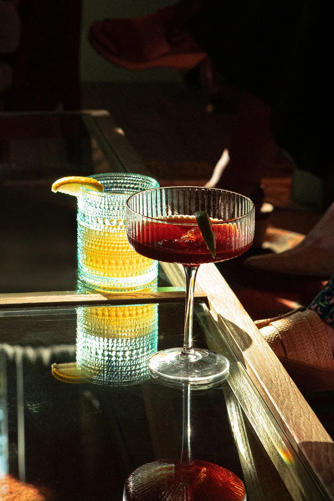 A cocktail glass with a reddish beverage and a lime wedge garnished on the side, placed on a reflective surface. Behind it, there is a beaded glass with a lemon wedge on the rim, also reflecting on the surface.