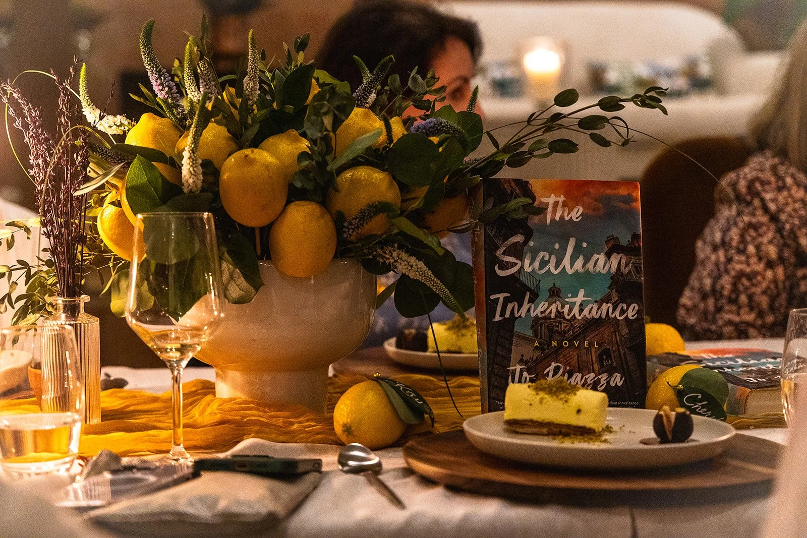 A table setting featuring a large bouquet of lemons, green leaves, and purple flowers in a white vase, a plate with a slice of lemon cheesecake, a book titled "The Sicilian Inheritance," some lemons, and empty wine glasses. In the background, there a