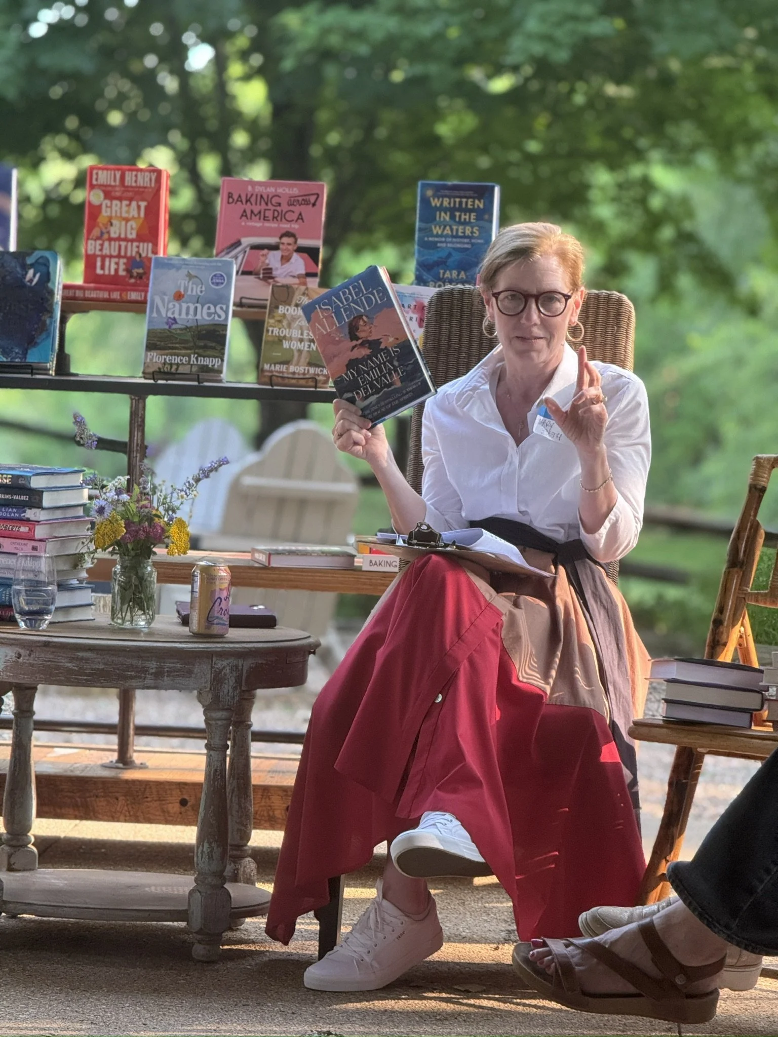 A woman with short hair and glasses sit on a wooden chair outdoors, holding a book titled 'My Name is Emilia Delavale' by Isabel Allende. She wears a white shirt and a long skirt with red, beige, and pink hues. Books are displayed on a table and shel