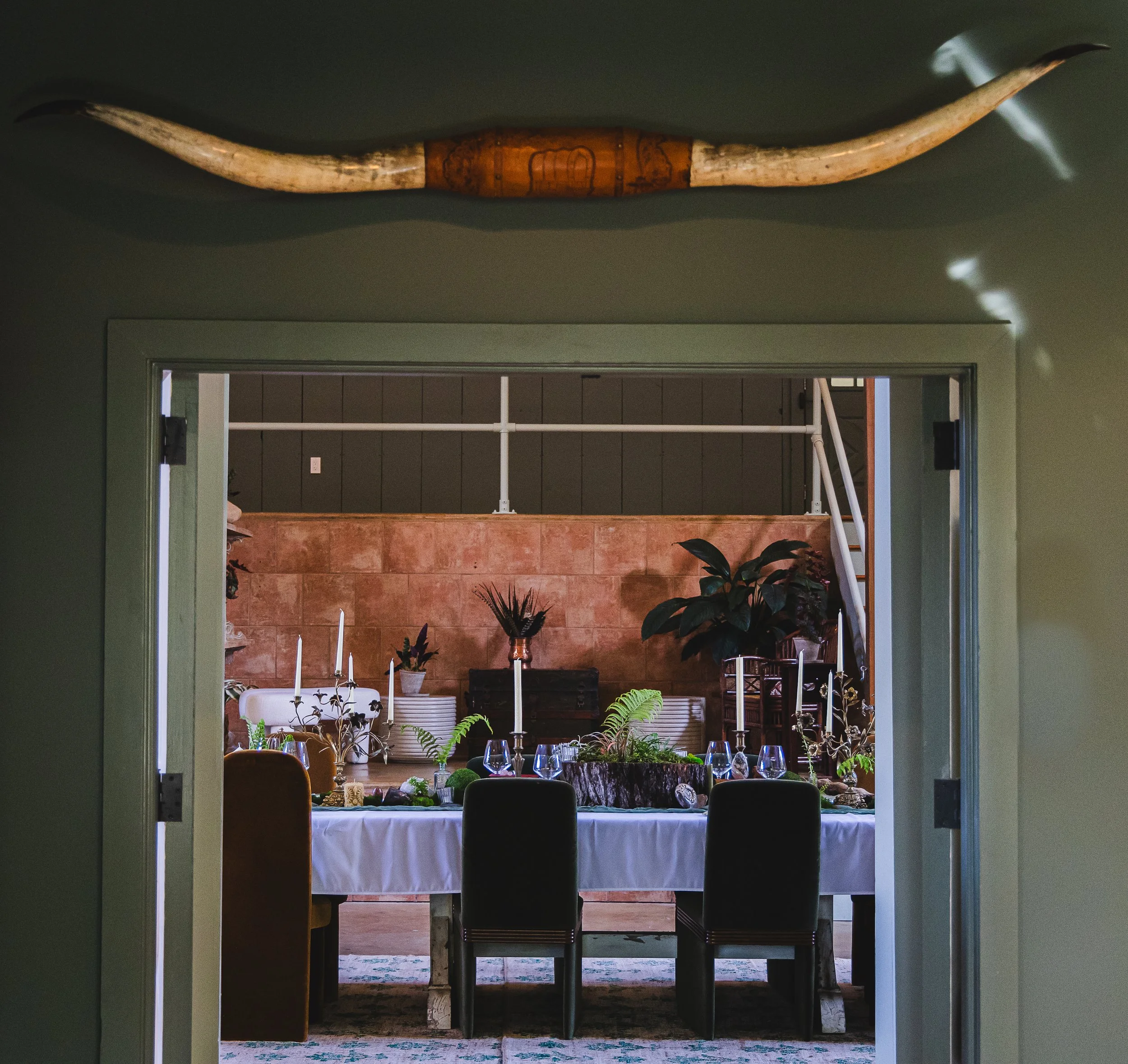 Decorated dining table with candles, glassware, and plants, viewed through a doorway with an animal horn mounted above.
