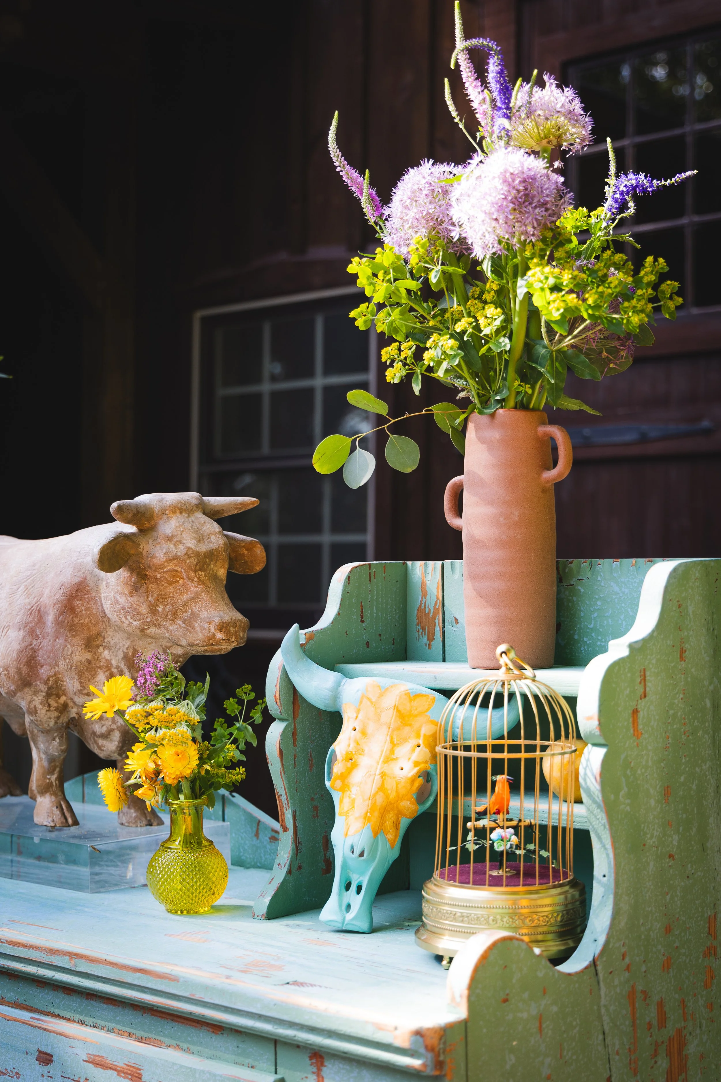 A rustic, distressed green wooden surface with decorative items, including a ceramic bull head, a small yellow vase with yellow flowers, a tall terracotta vase with pink and purple flowers, a small bird in a golden cage, and a decorative yellow leaf.
