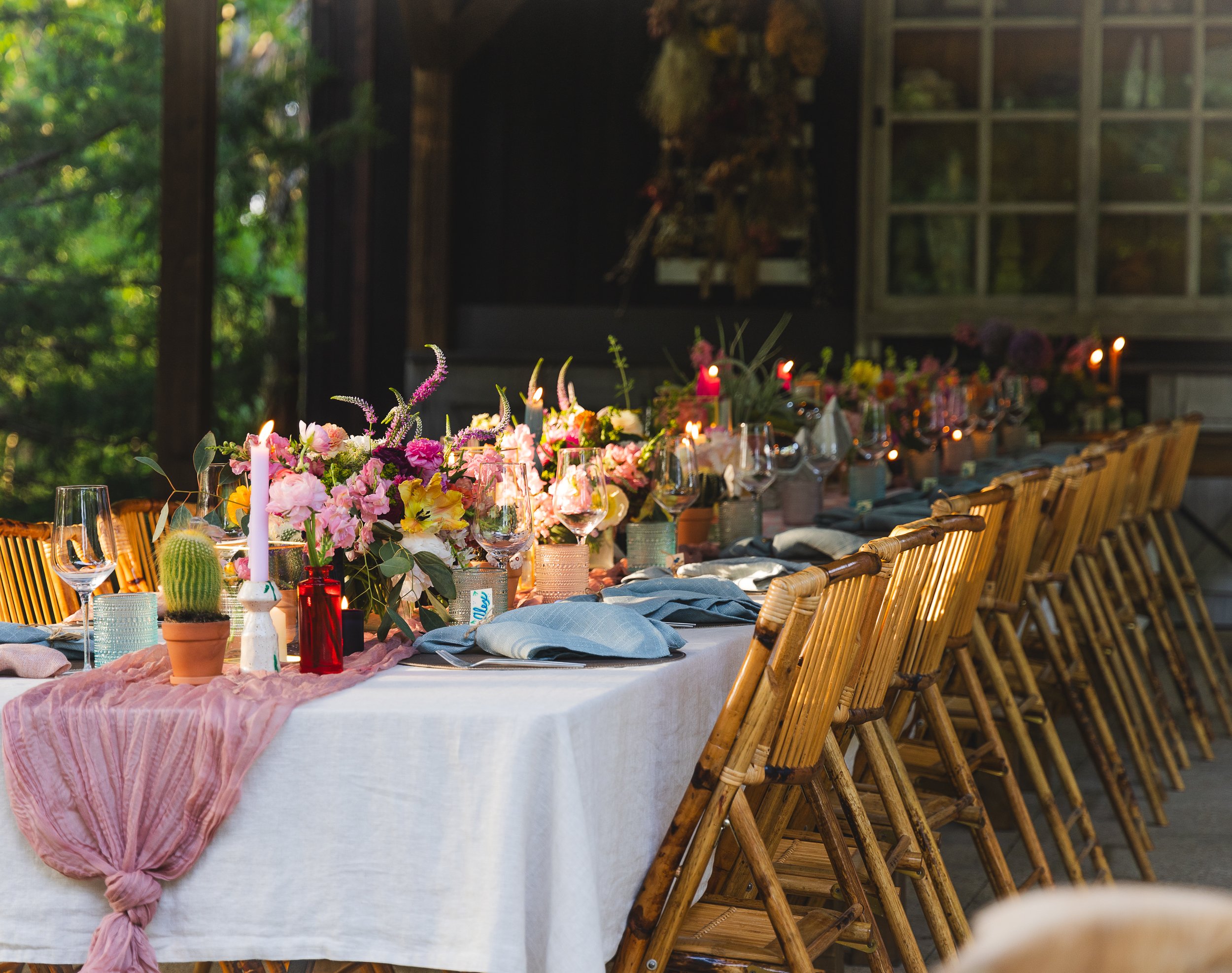 Wedding or event reception table decorated with pink and purple flowers, candles, and tableware, with chairs on one side and a rustic wooden building in the background.