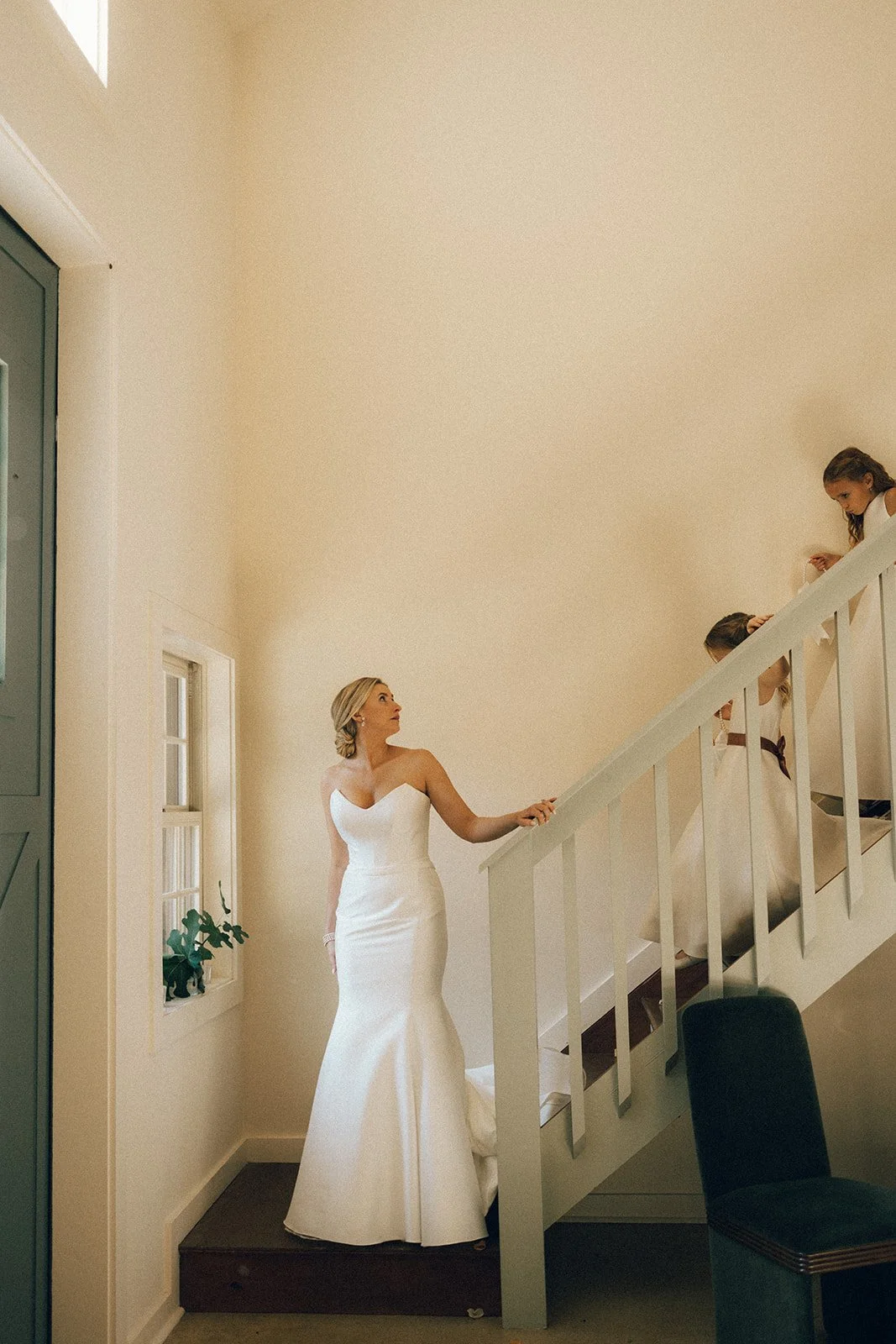 A bride in a white wedding dress standing at the bottom of a staircase, looking up at three young girls on the staircase.