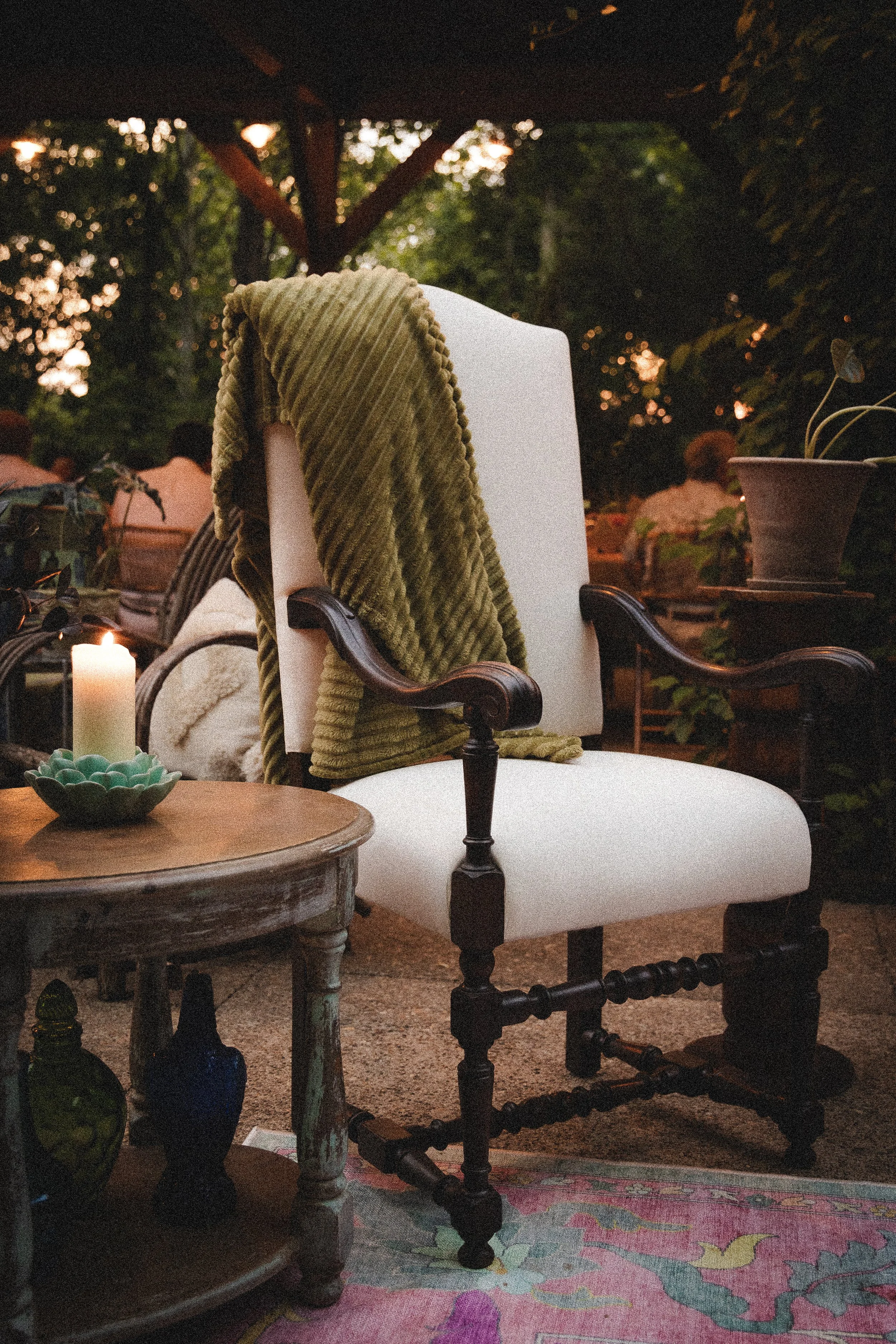 A cozy outdoor seating area featuring a white upholstered antique wooden armchair with dark wooden arms, draped with a green textured blanket. A small wooden side table with decorative bottles and a white candle is in front of the chair, set on a pat