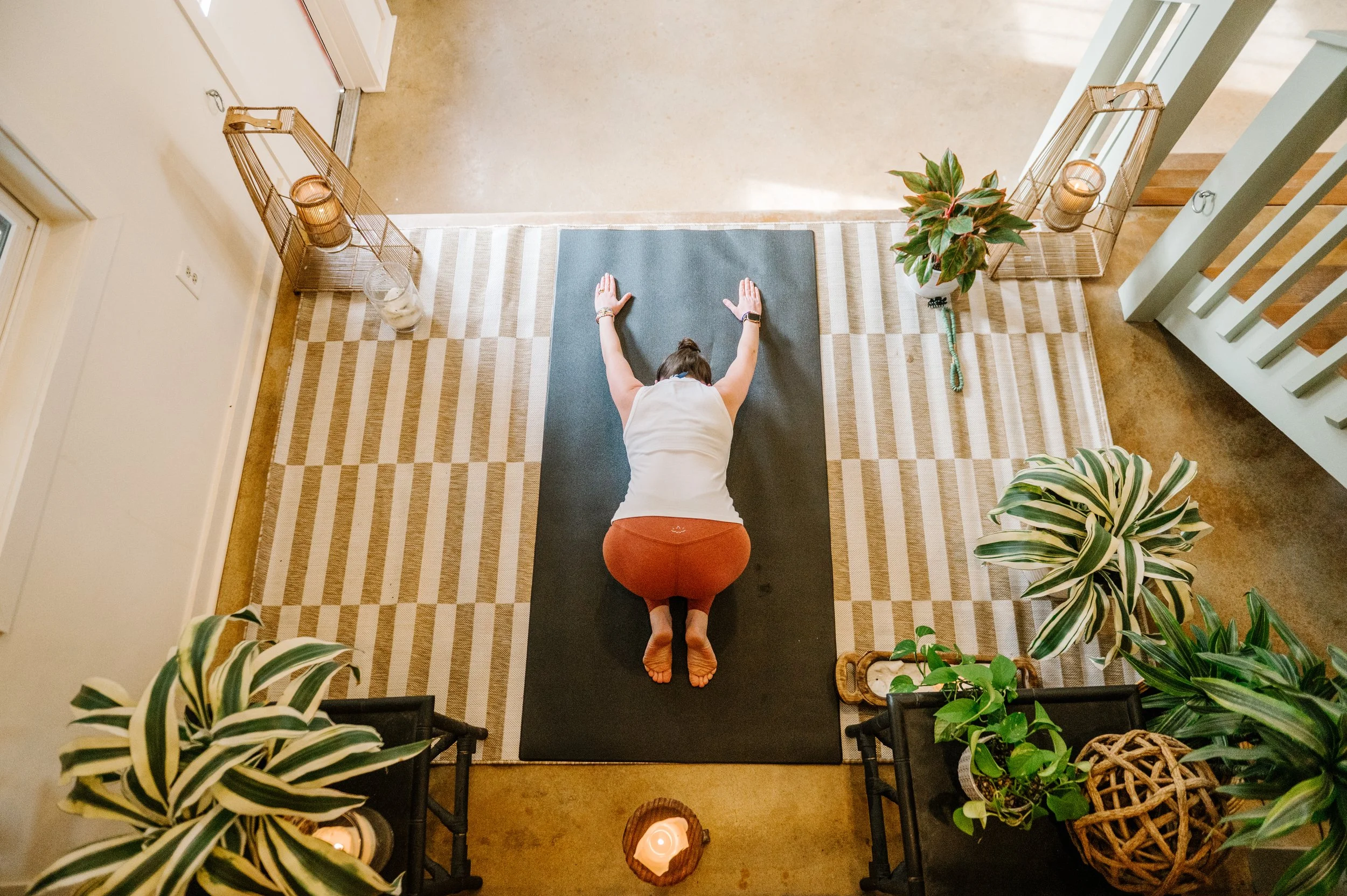 A woman practicing yoga on a black mat in a cozy indoor setting, surrounded by green plants and soft lighting.