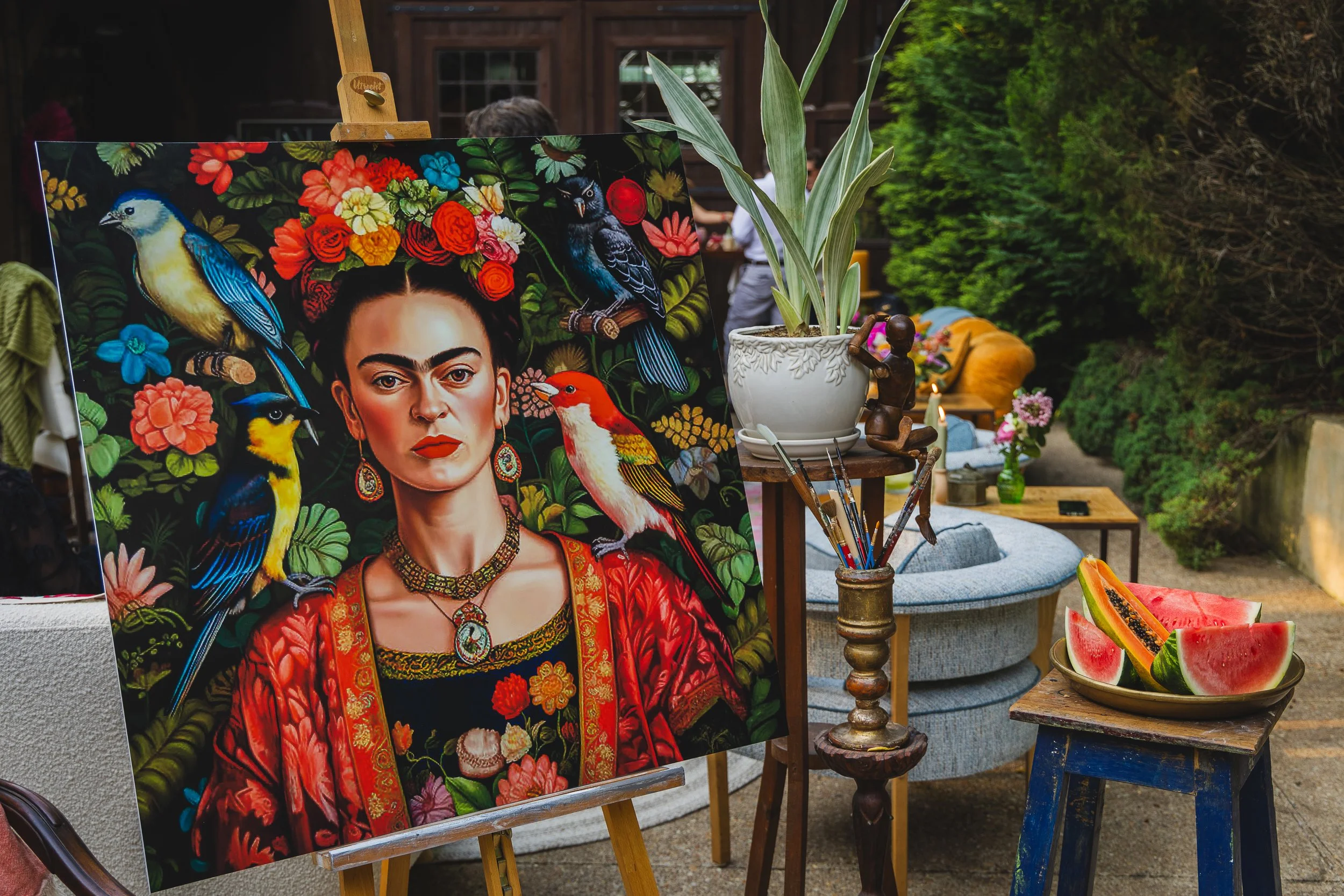 A colorful portrait painting of Frida Kahlo surrounded by various birds, displayed on an easel at an outdoor art viewing area. Watermelon slices in a bowl are in the foreground, and a potted plant along with other art supplies and furniture are visib