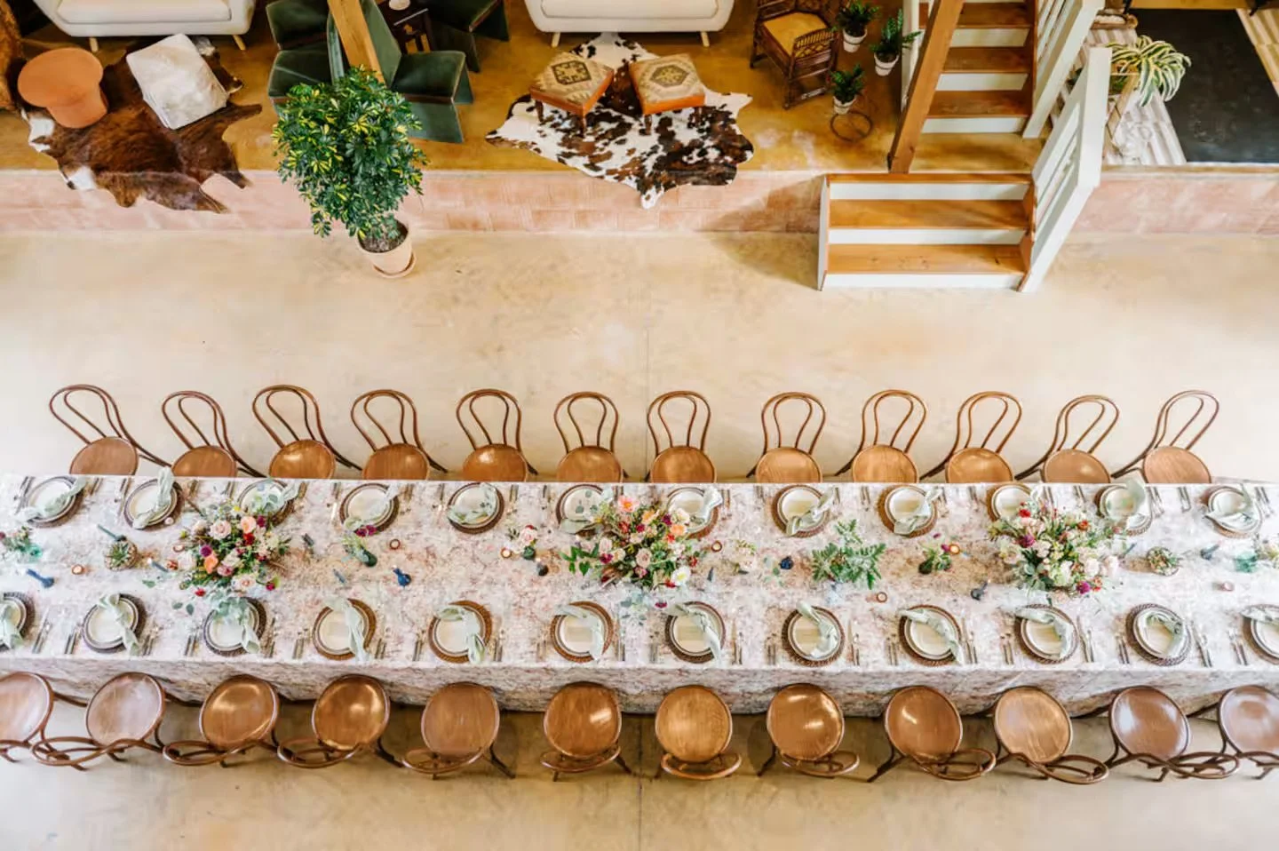 A long dining table set for a meal with floral centerpieces, plates, glasses, and utensils. Surrounding the table are wooden chairs with curved backs. The room features wooden stairs, a cowhide rug, a potted plant, and additional seating in the backg