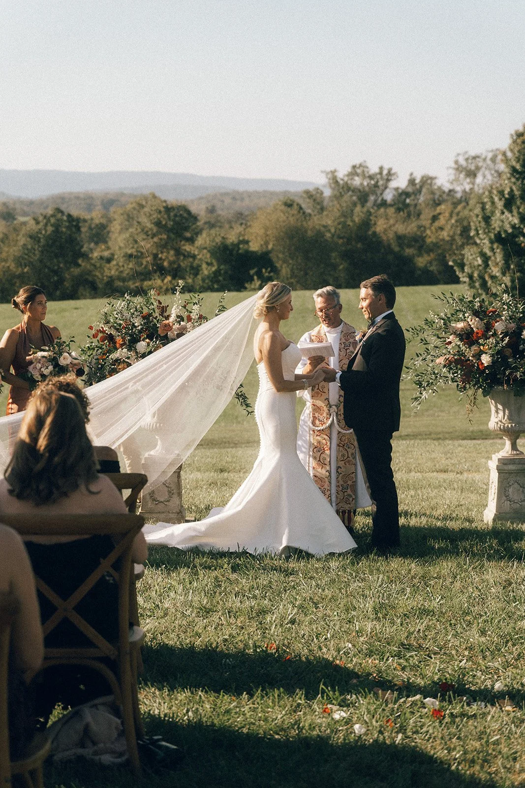 Bride and groom exchanging vows during outdoor wedding ceremony, officiant present, guests seated, scenic background.