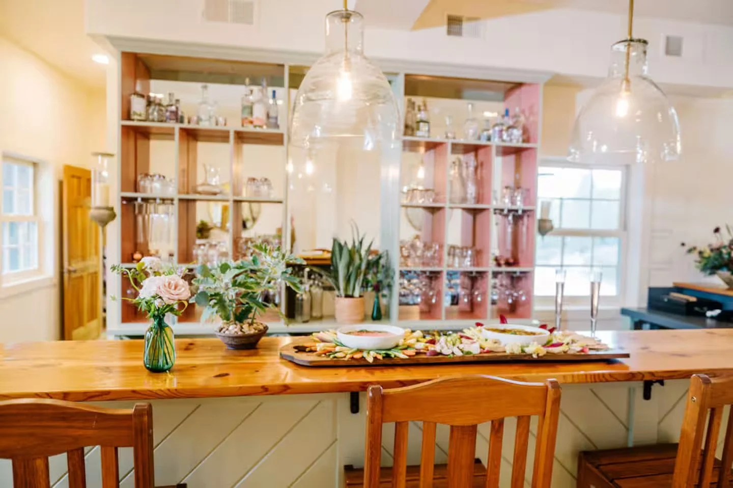 A wooden kitchen island with flower arrangements and bowls on top, with open shelving and glassware in the background, and pendant lights above.